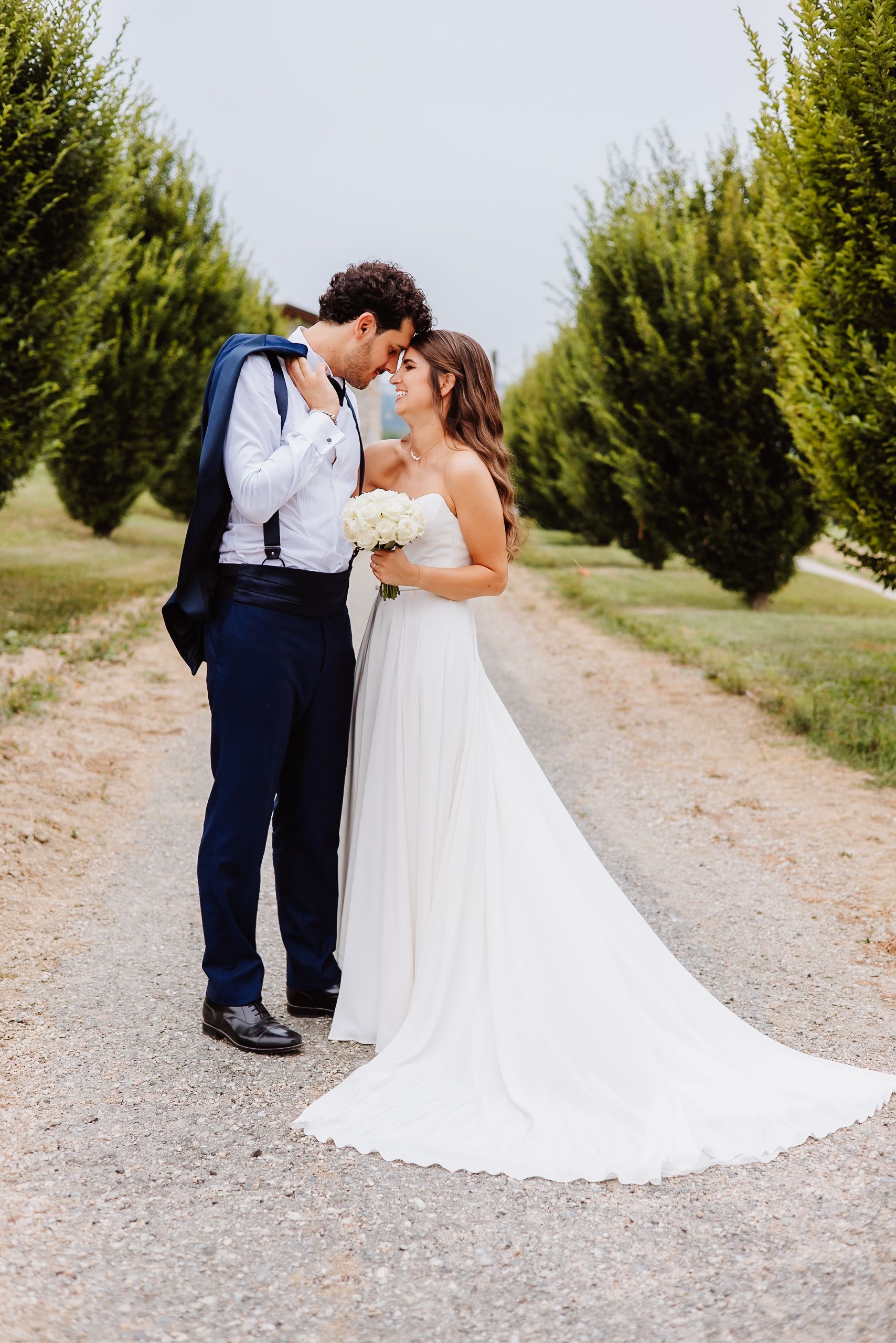 A bride and groom share a romantic moment on a gravel path lined with cypress trees on their wedding day.