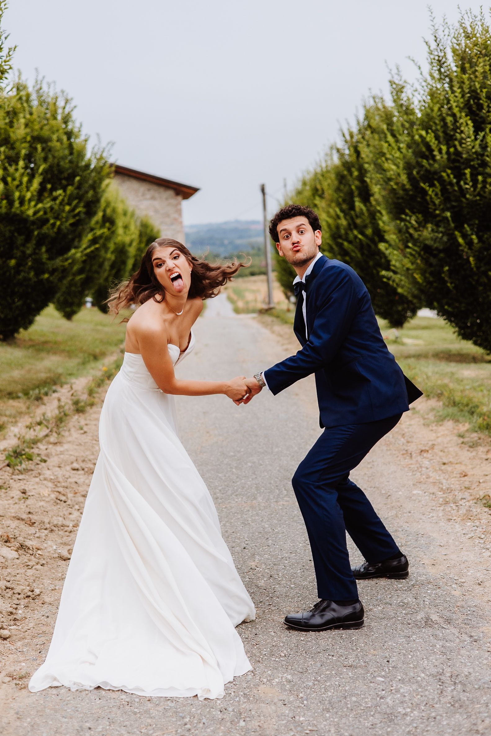 A couple in wedding attire playfully pose for a fun photo sequence on a dirt path surrounded by greenery.
