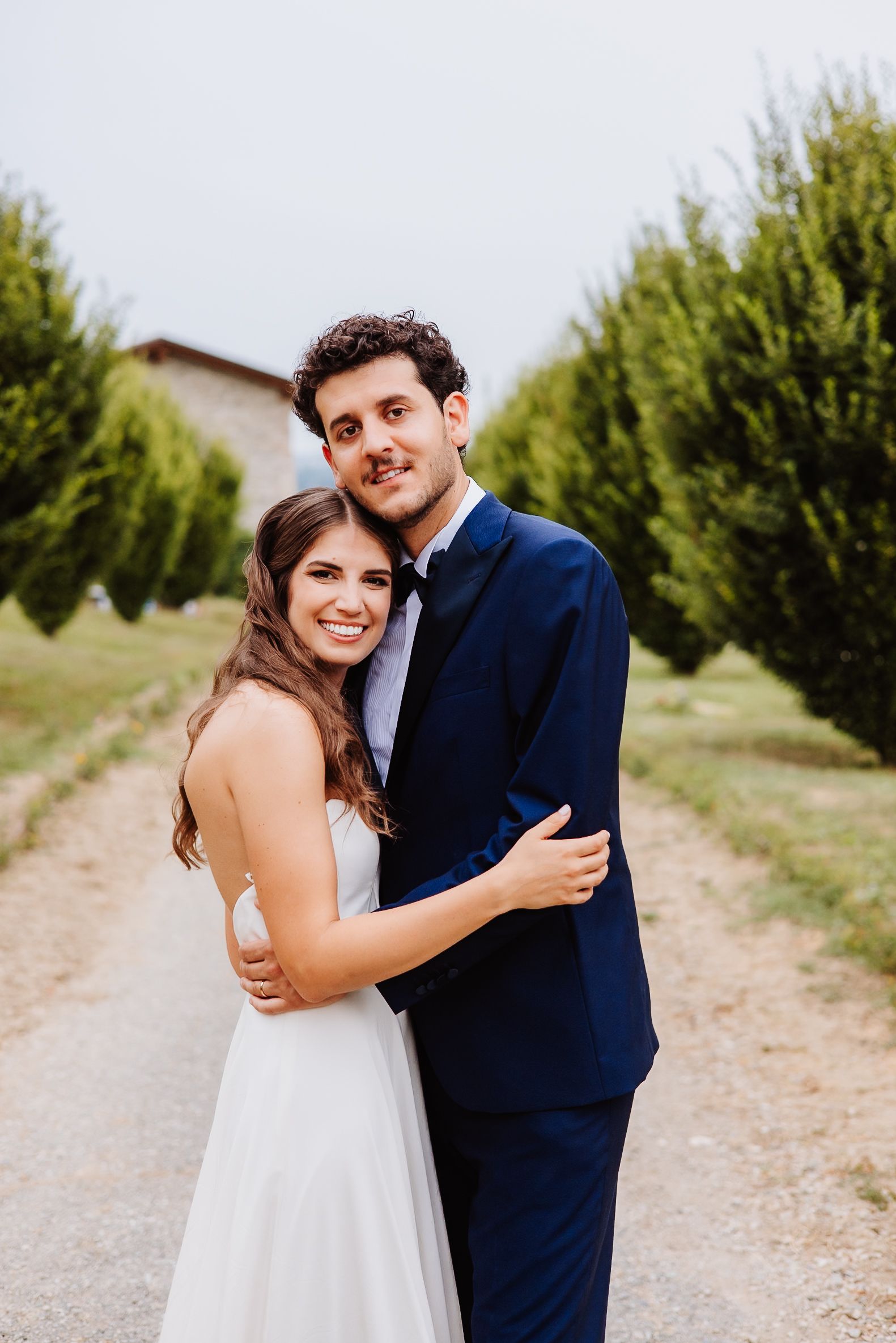 A happy couple laughs together during their outdoor wedding photos along a gravel path lined with green trees.