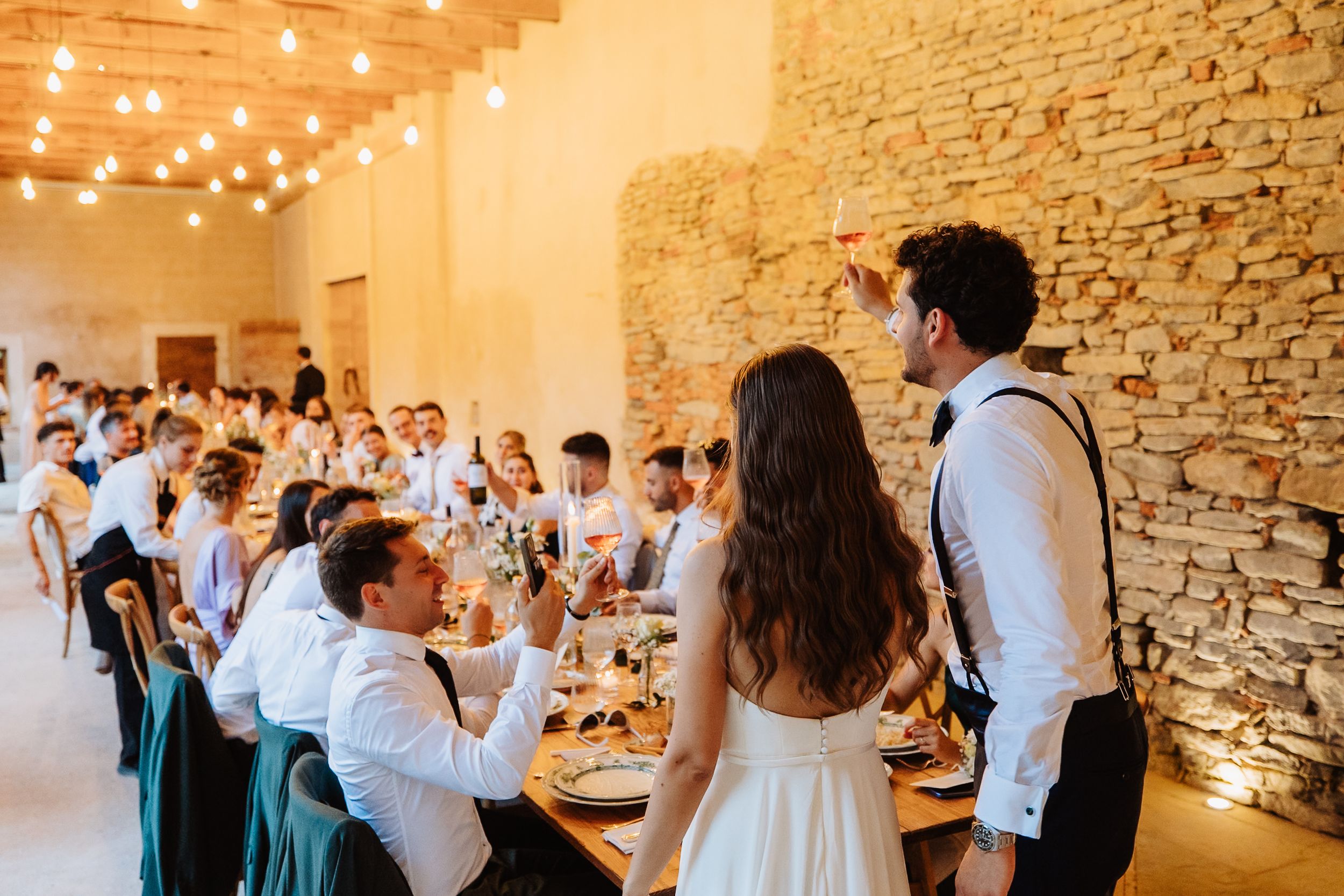 Wedding party dancing and celebrating in rustic venue with stone walls and string lights overhead during reception.