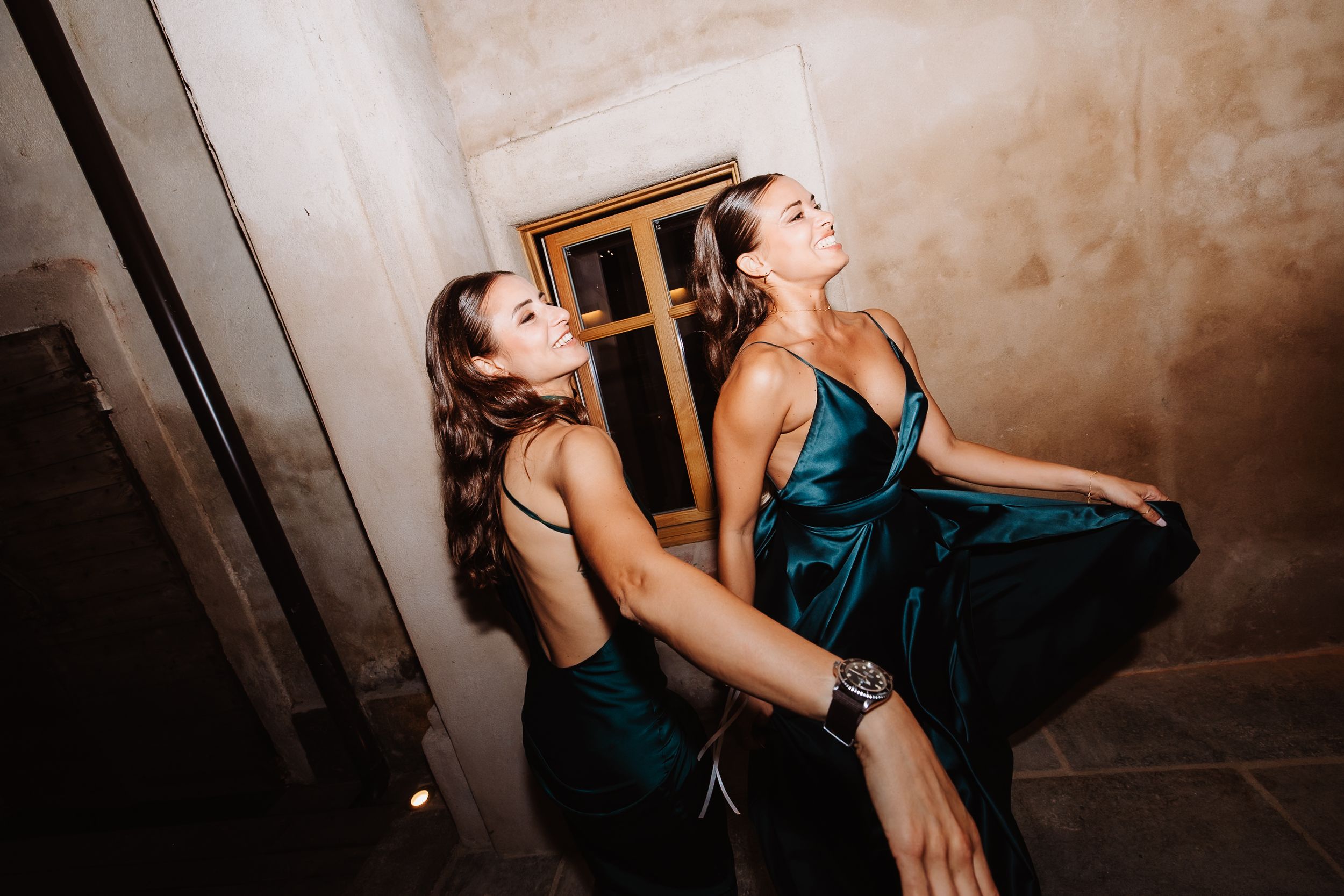 Two women in emerald satin dresses laughing and dancing together at a party against a textured wall.