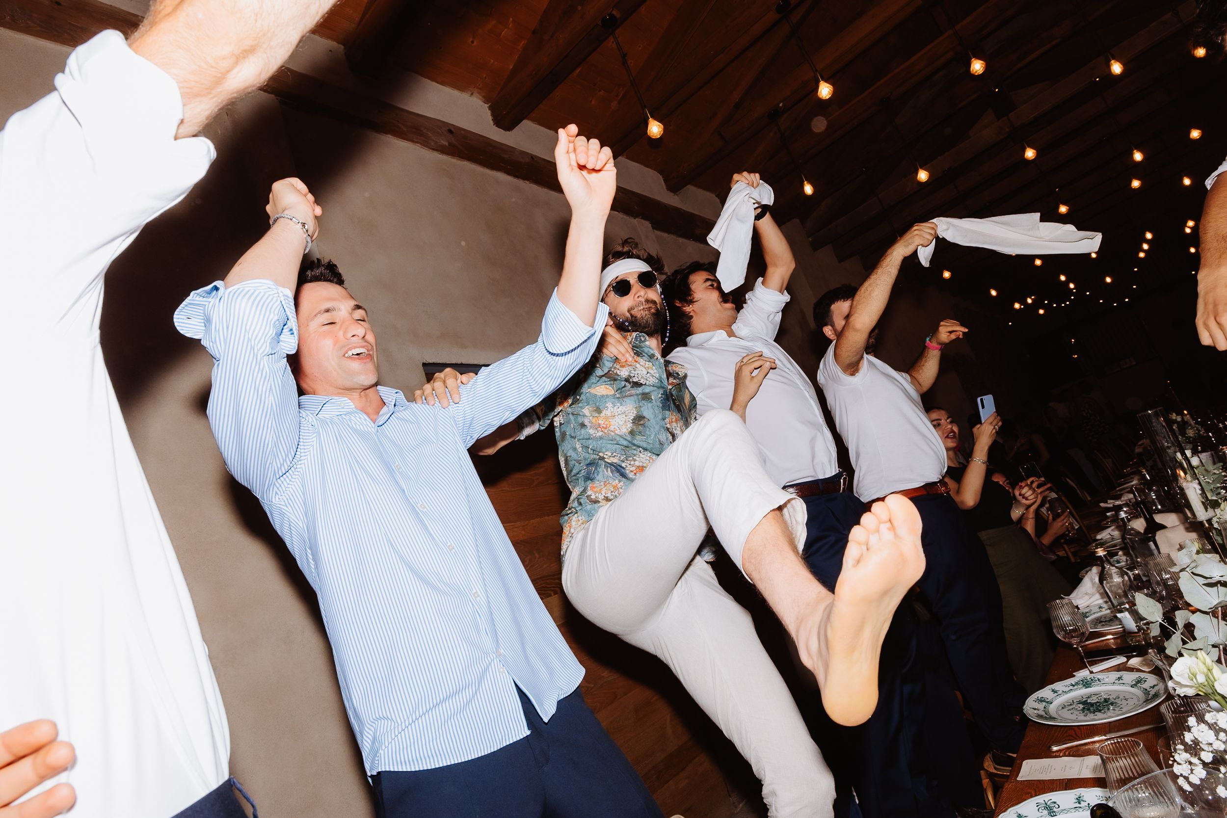 People dance enthusiastically in a dimly lit reception hall with strings of lights visible in the background.