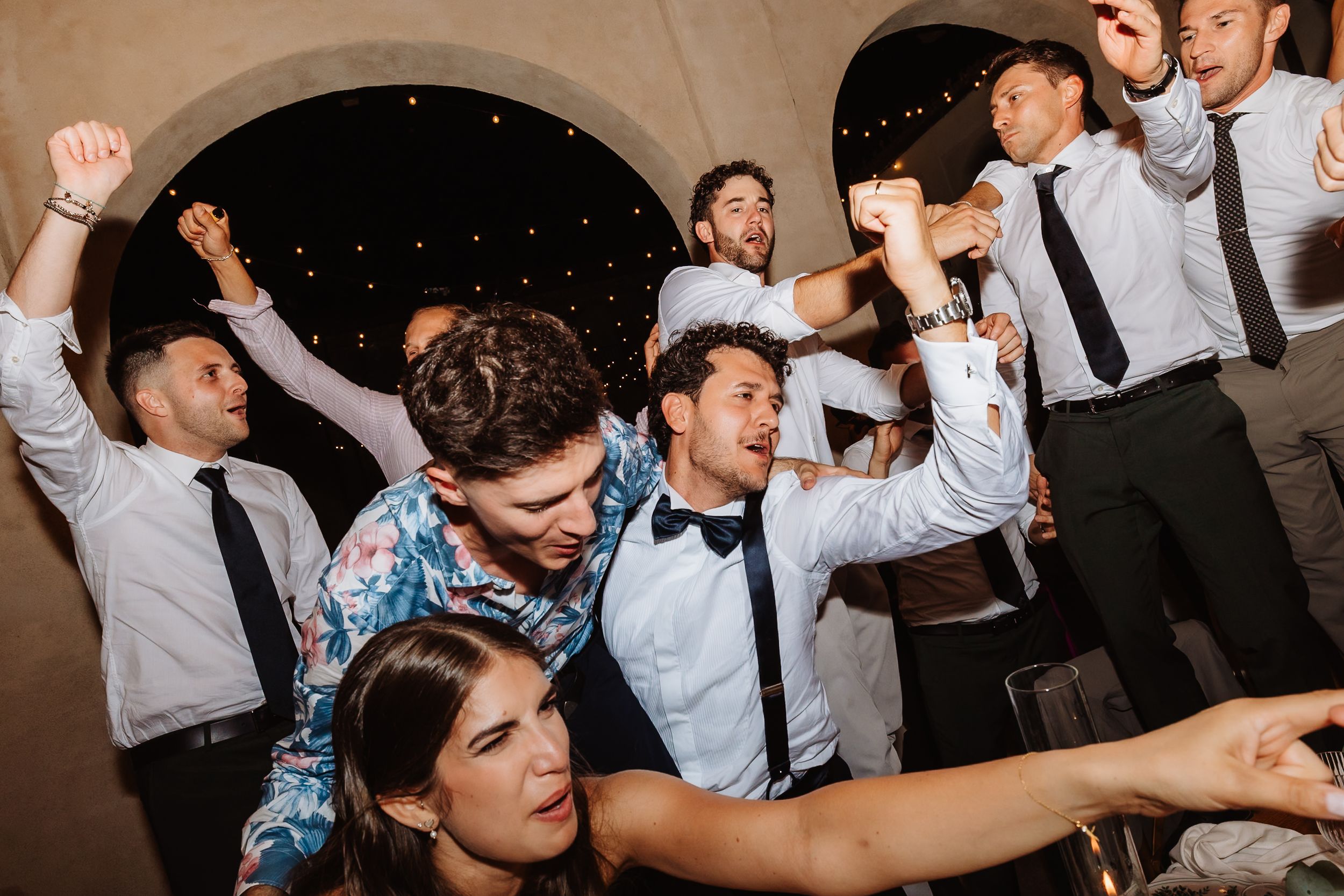 A group of party guests in formal attire dancing and celebrating energetically at a wedding reception.