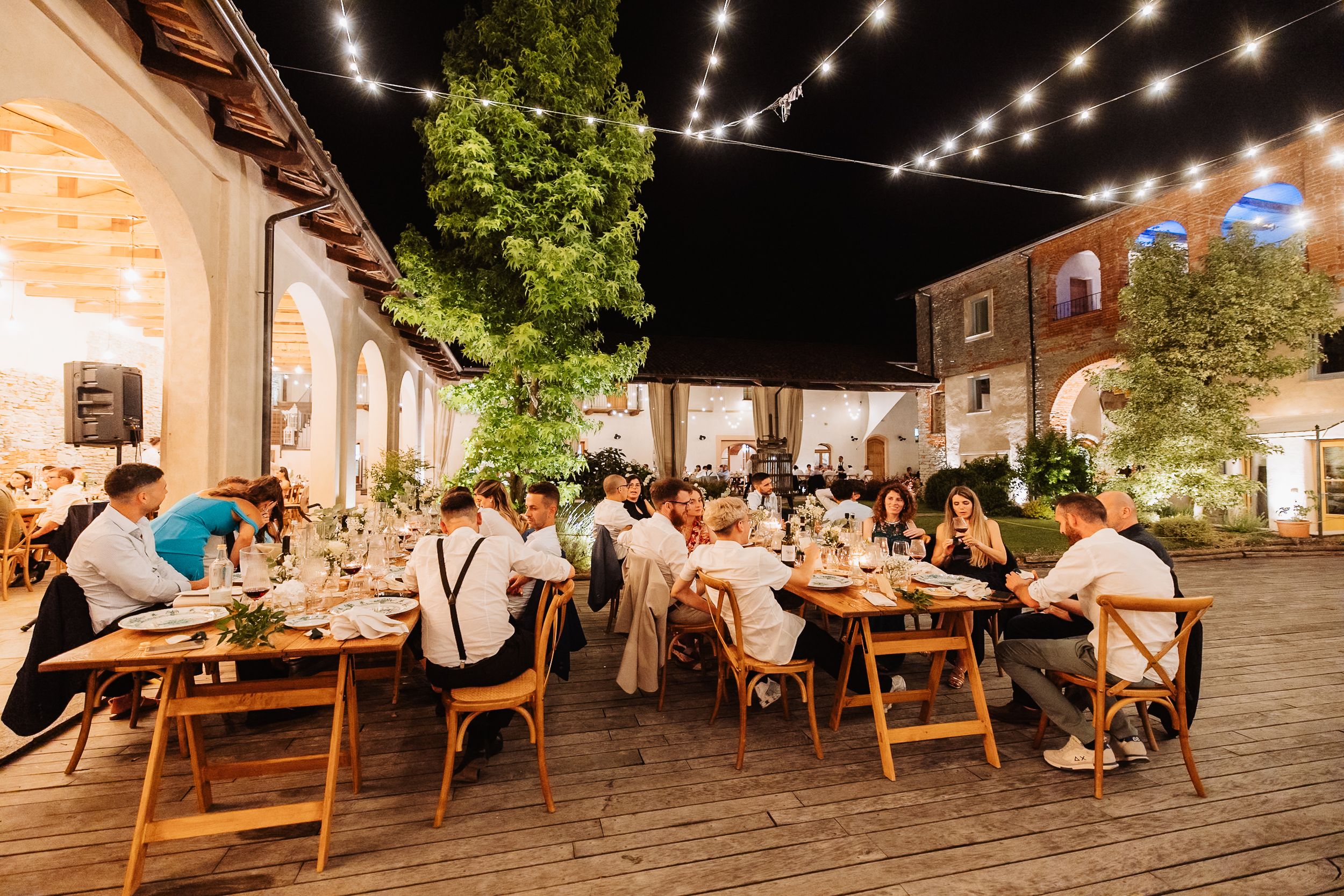 String lights illuminate an outdoor dining patio with wooden picnic tables and guests enjoying evening meals.