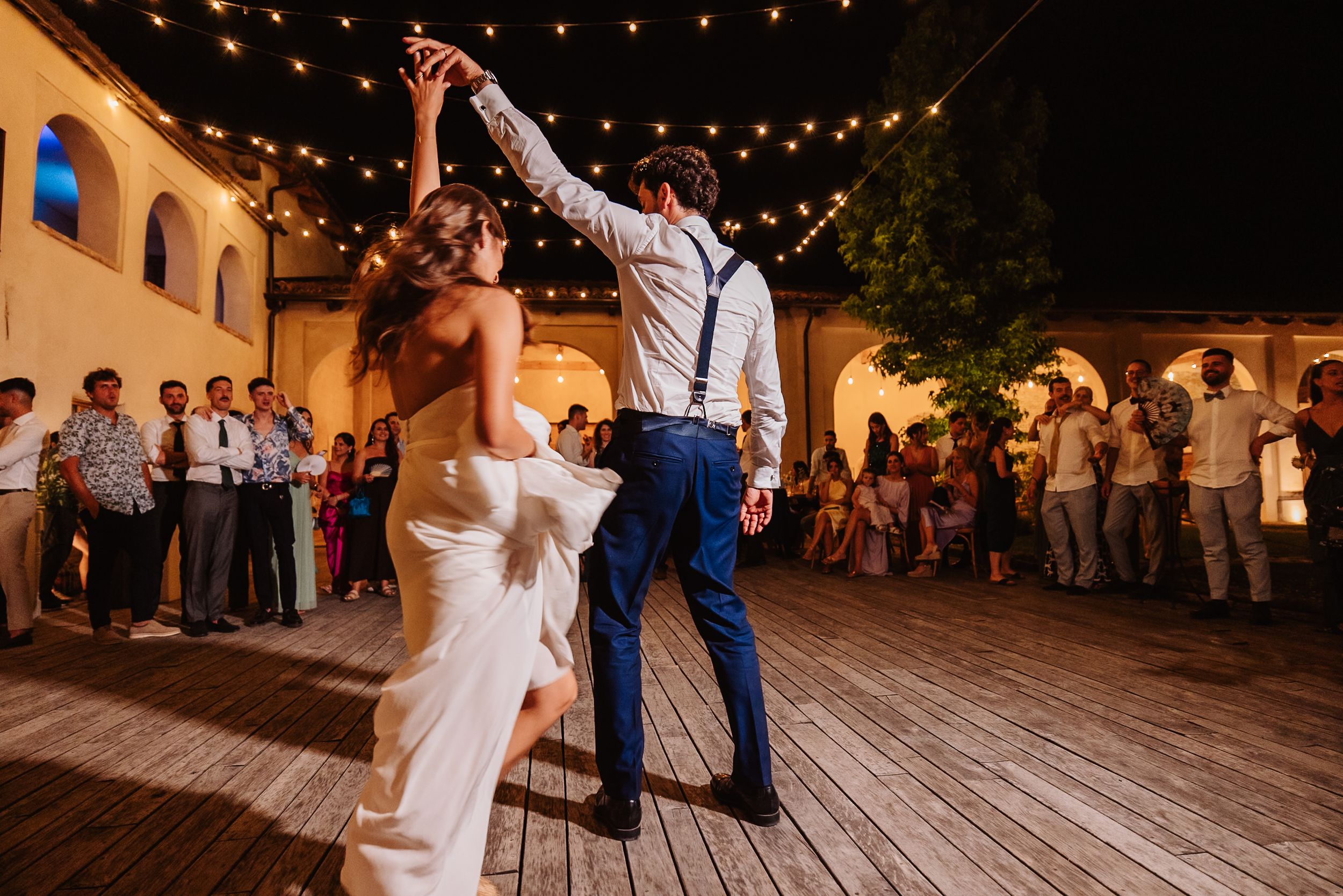 A couple dances together under romantic string lights during an evening outdoor reception celebration.