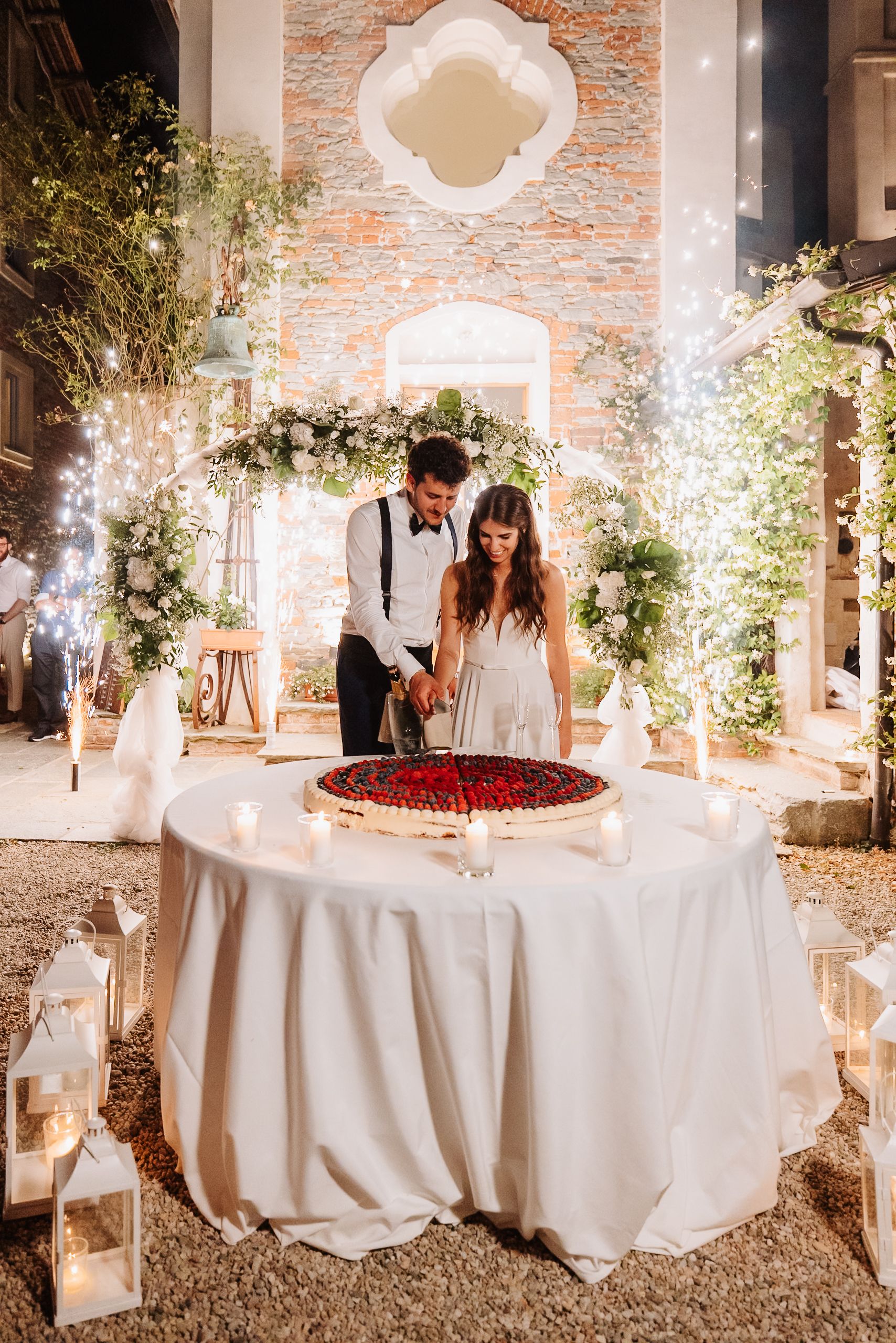 A romantic wedding scene with a beautifully decorated cake table featuring sparklers and floral arch in a rustic brick venue.