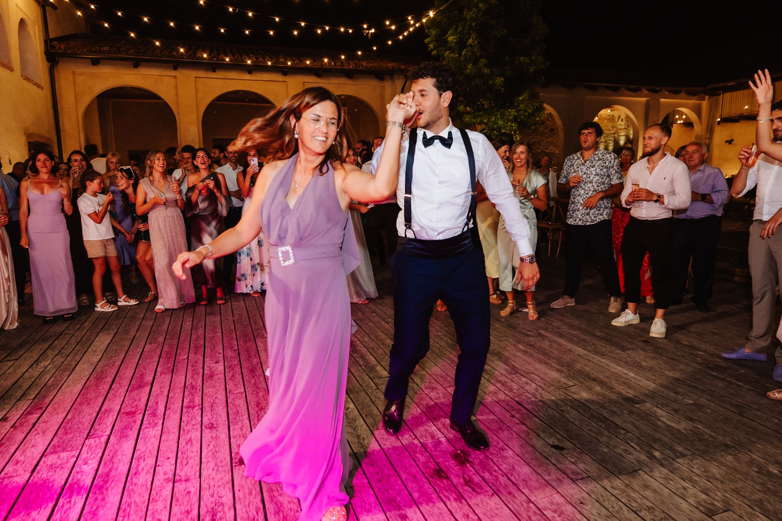 Two people dance together on a wooden deck illuminated by pink lighting and overhead string lights at an evening celebration.