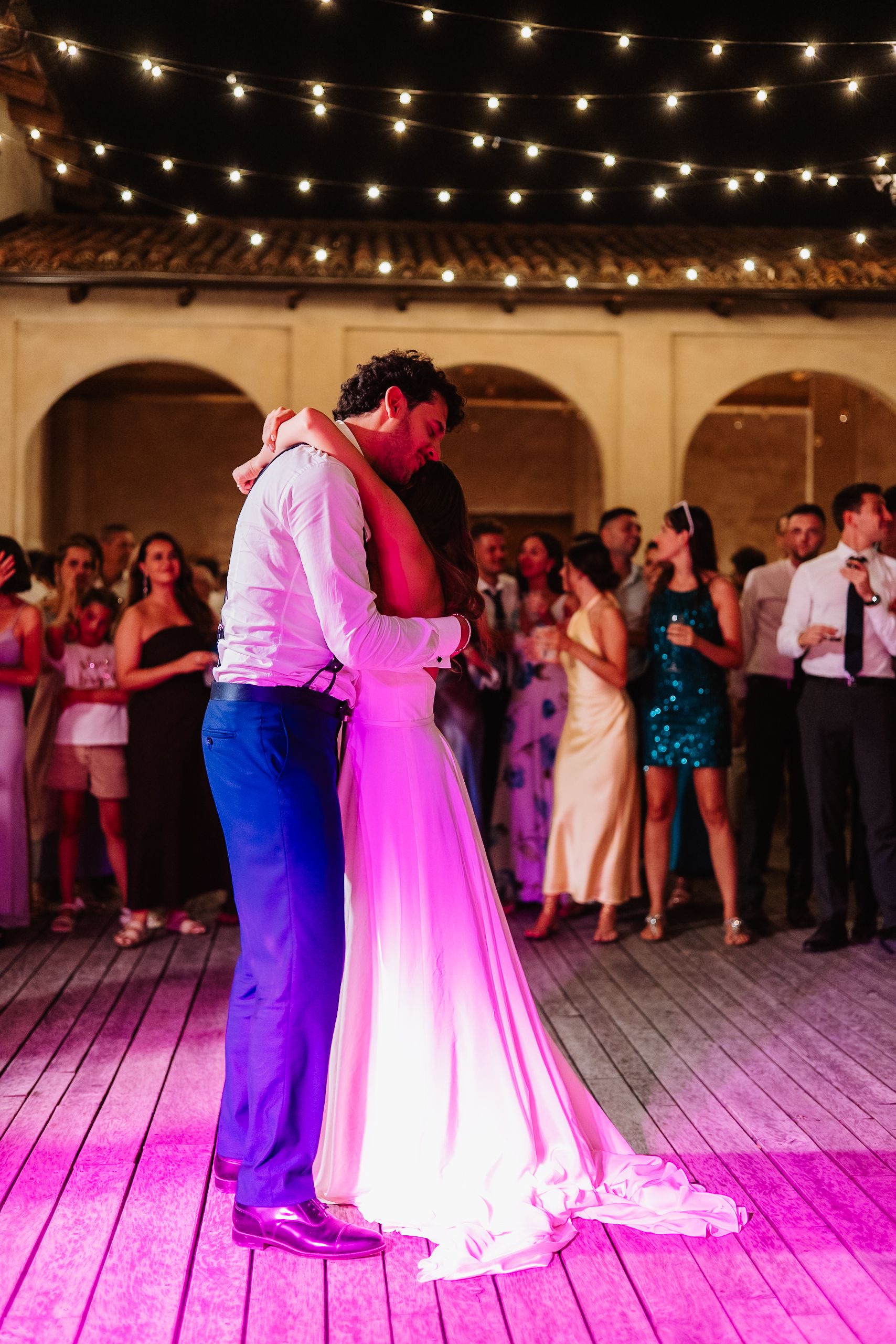 A couple shares their first dance at a wedding reception under string lights while guests watch from the dance floor.