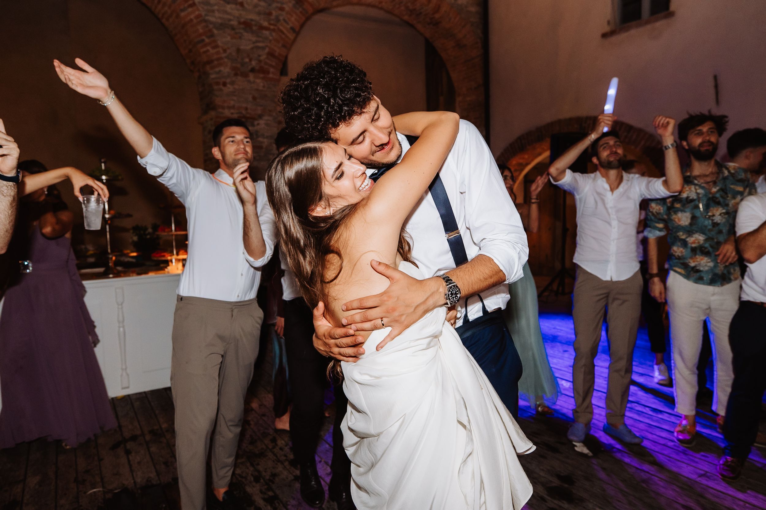 Couple shares romantic dip kiss on illuminated dance floor while wedding guests celebrate in background.