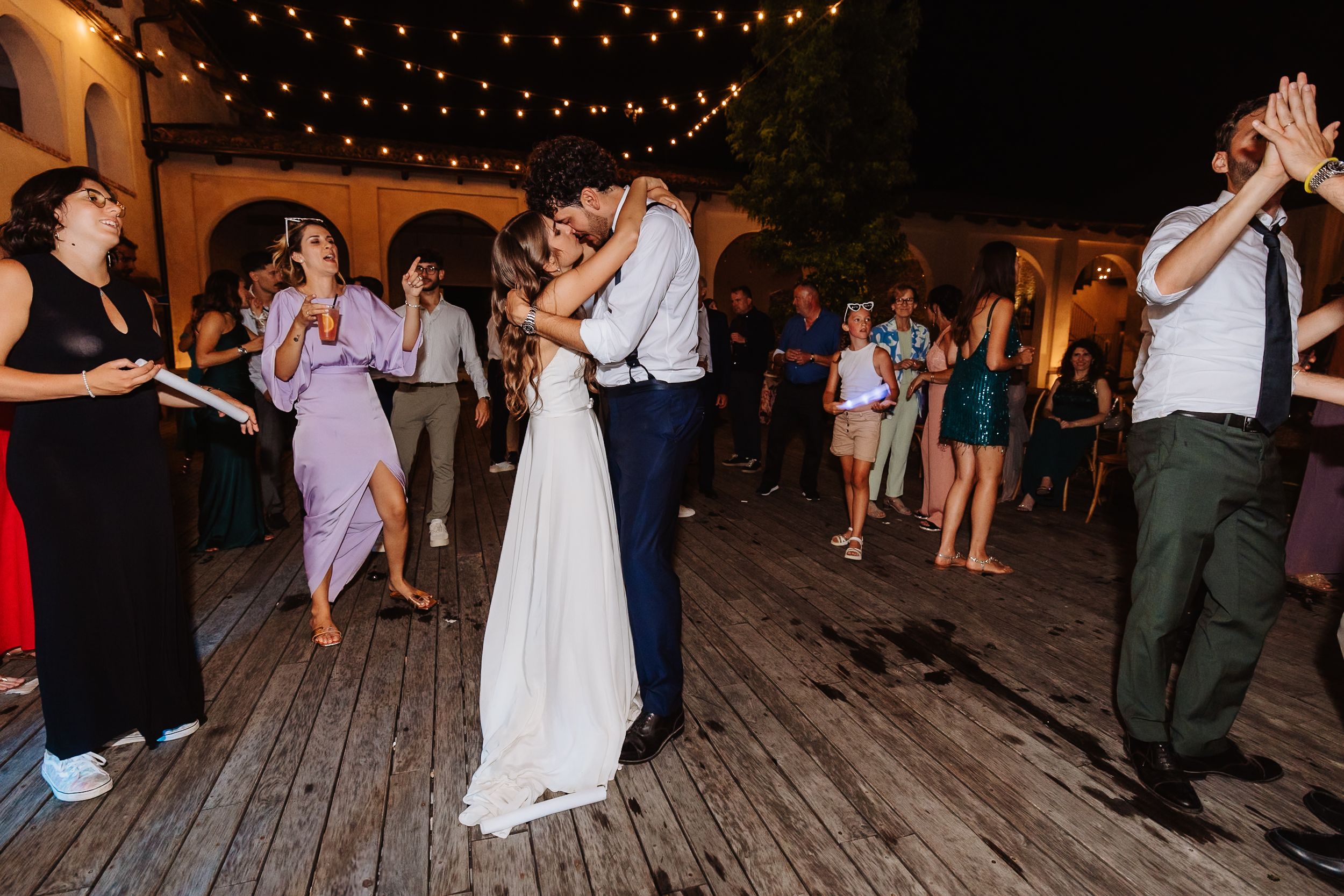 Couples dancing on a wooden deck under twinkling string lights at an outdoor evening reception.