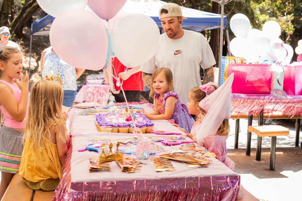 A festive pink outdoor party setup with balloons, tablecloths, decorations and people gathered around tables.