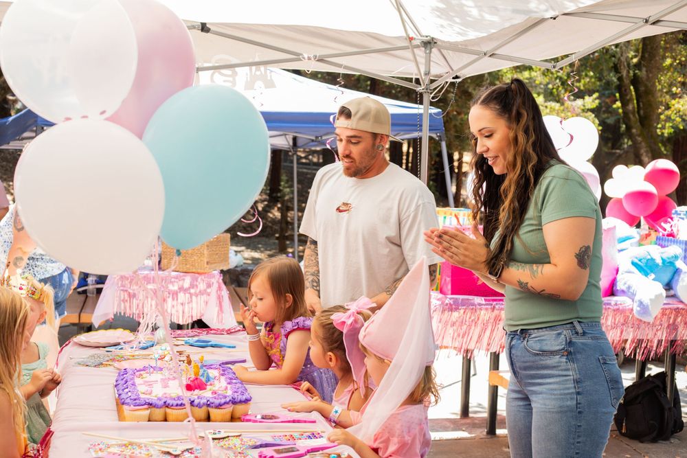 Pink and white party decorations with balloons adorn tables at an outdoor event under white canopy tents.