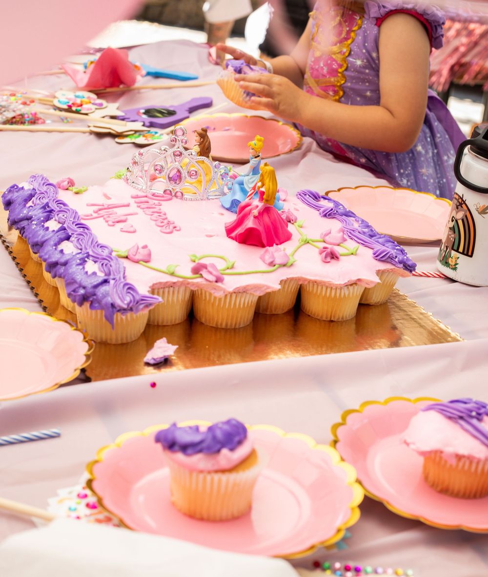 Close-up view of cupcakes with pink and purple frosting being decorated on pink plates at a party activity table.