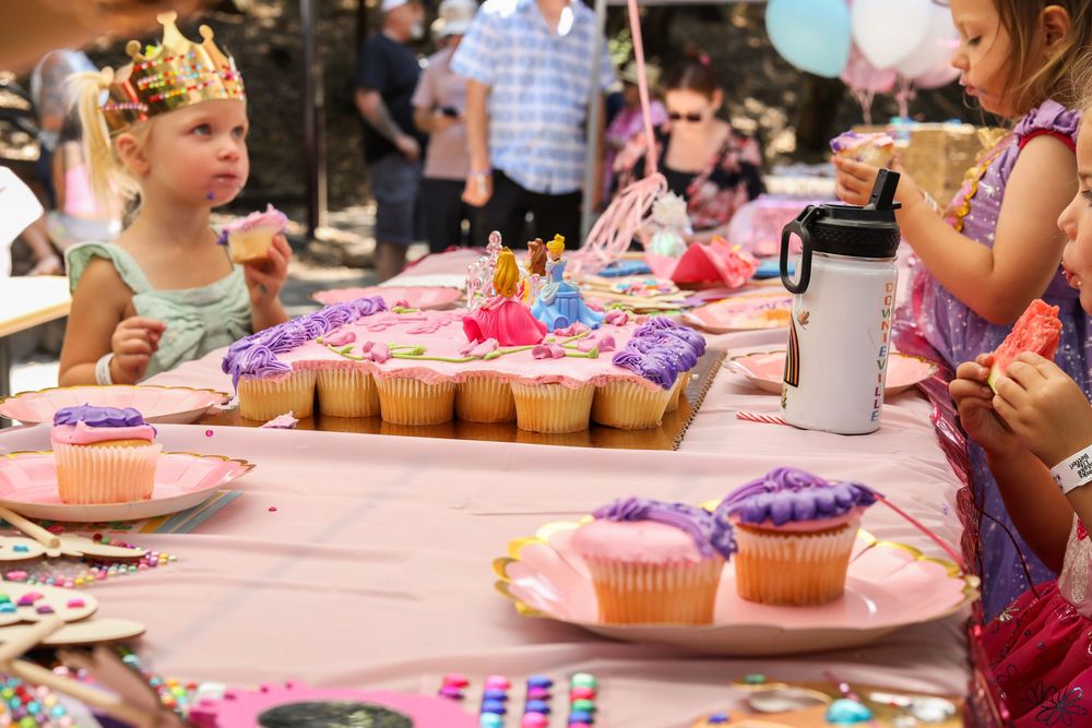 Children decorate colorful cupcakes with frosting and sprinkles at a party table covered in pink tablecloth.