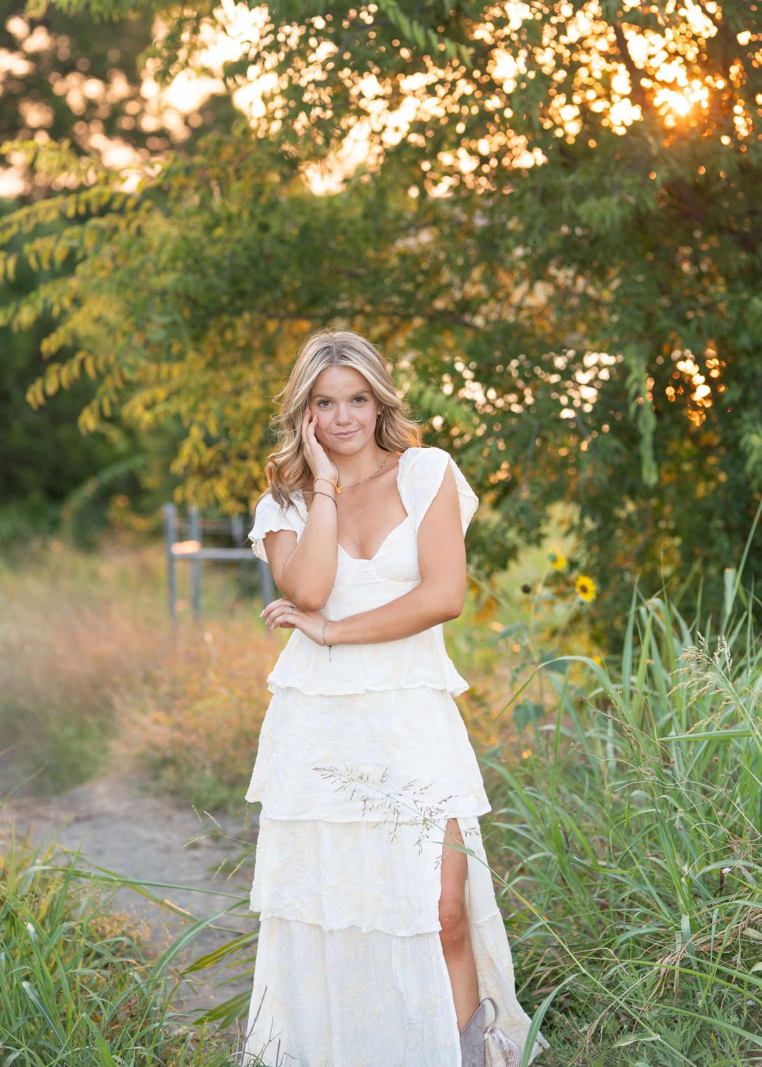 A figure in a flowing white dress poses gracefully in tall grass during golden hour sunset.