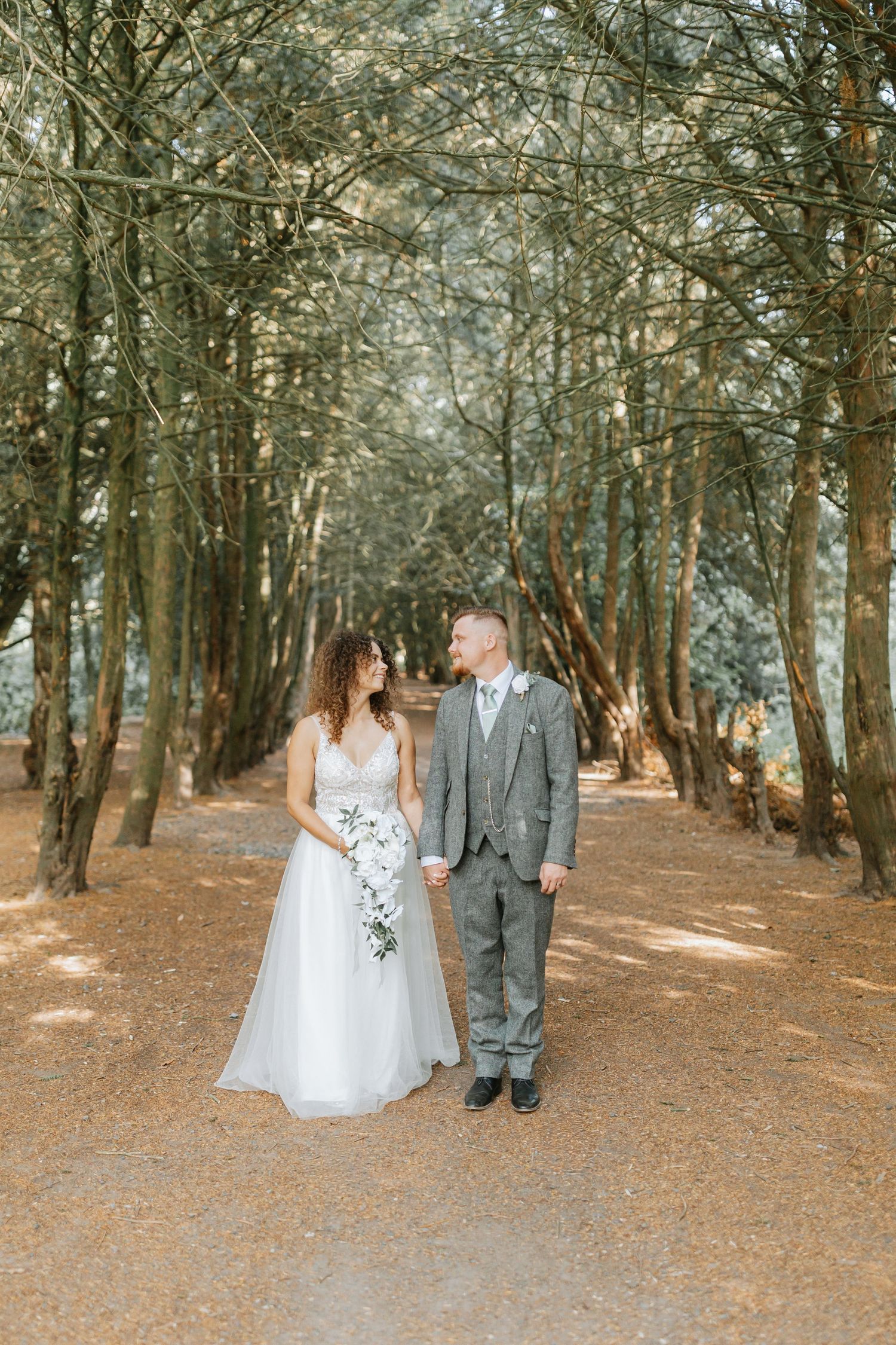Wedding couple walks hand in hand through a romantic path lined with tall pine trees in an enchanting forest setting.