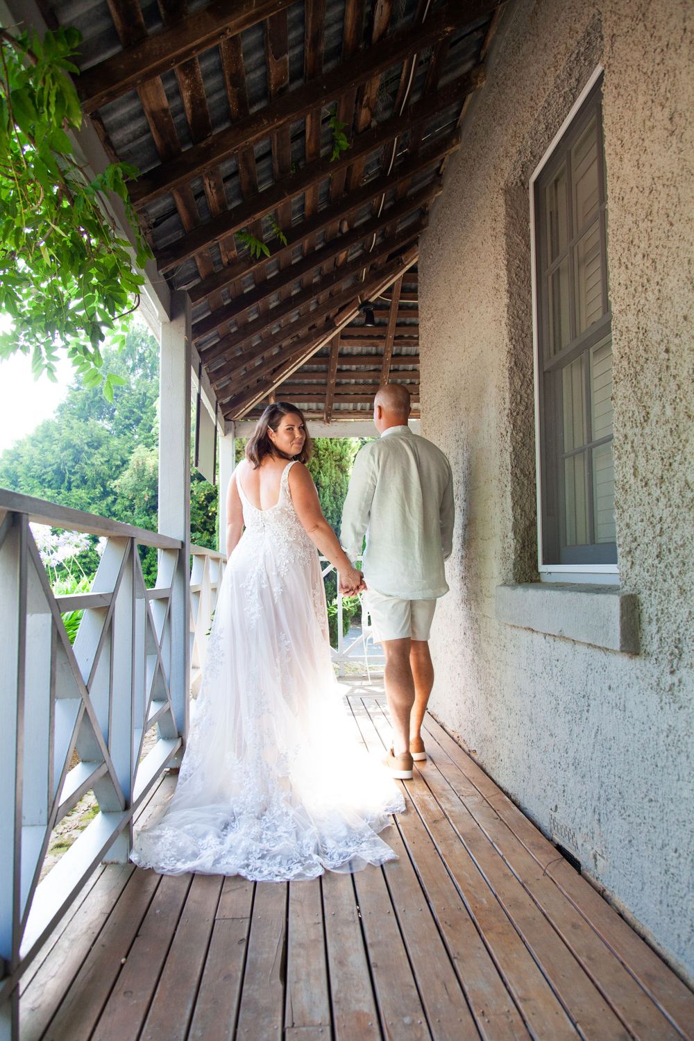 A couple in white attire walking along a rustic covered porch with wooden floorboards and trailing lace dress detail.