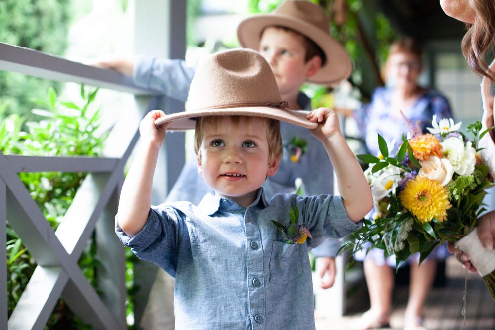 A young child in denim and a light brown hat stands among flowers at an outdoor event.