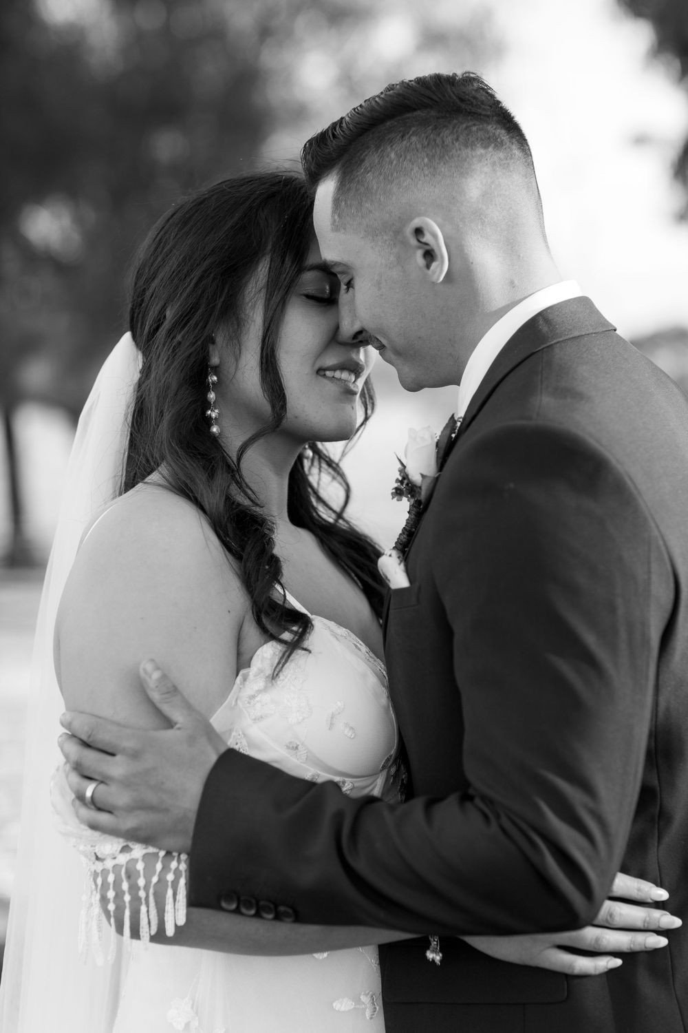 A romantic black and white wedding portrait shows a tender moment between a bride and groom embracing outdoors.