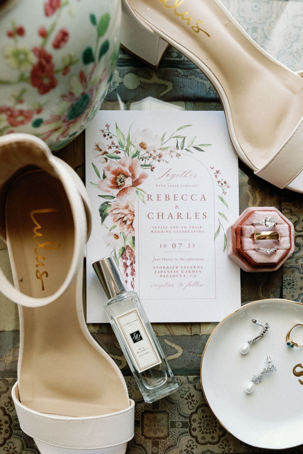 Flat lay of wedding invitation for Rebecca and Charles with blush heels, jewelry, and perfume.