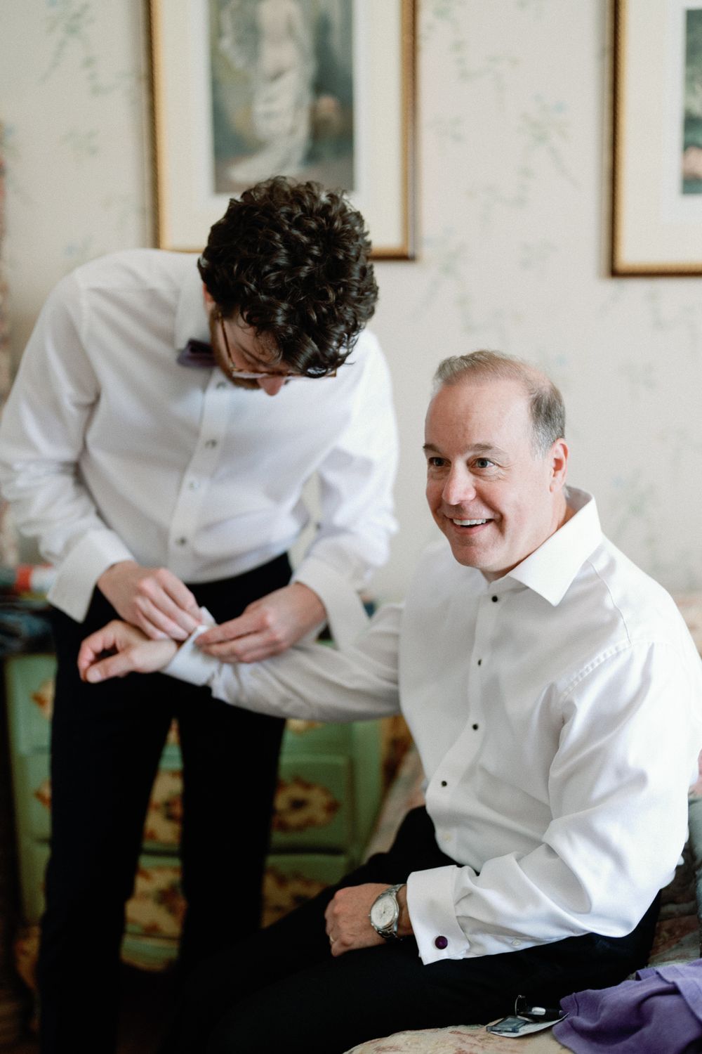 Smiling older man in white dress shirt getting cufflinks fastened by younger man while seated.