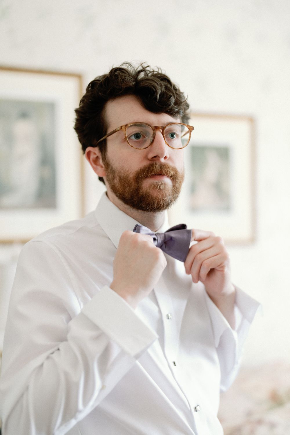 Bearded groom in white shirt and glasses adjusting purple bow tie while getting ready for wedding.