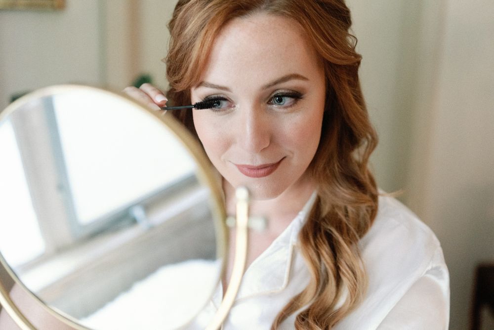 Beautiful red-haired bride applying mascara while looking into a round mirror during wedding prep.