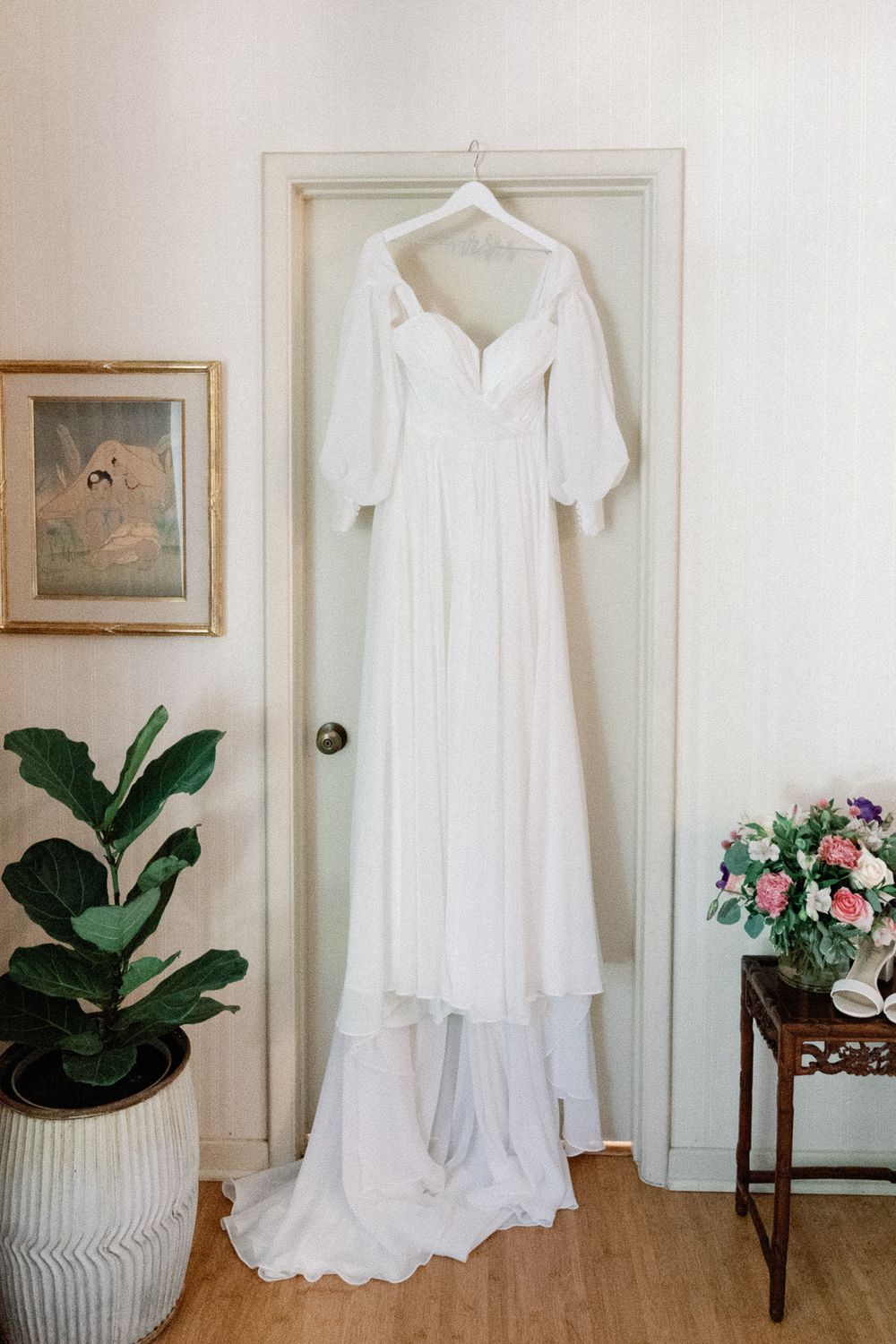 Elegant white off-shoulder wedding gown with puff sleeves hanging on a door in a bright room.