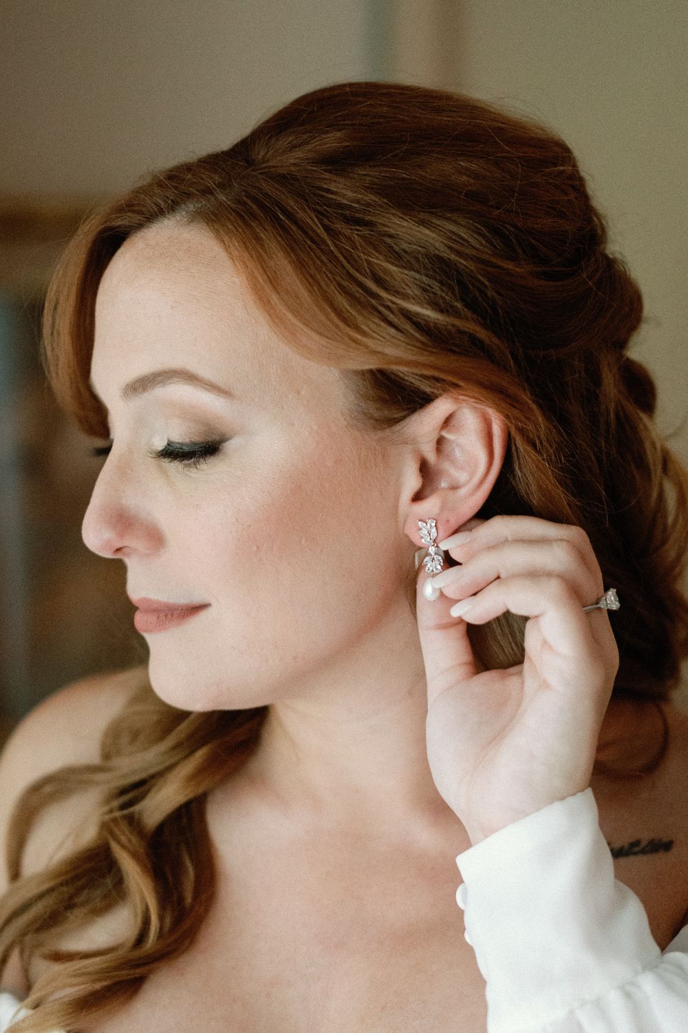 Close-up of red-haired bride putting on sparkling crystal drop earrings during wedding preparation.