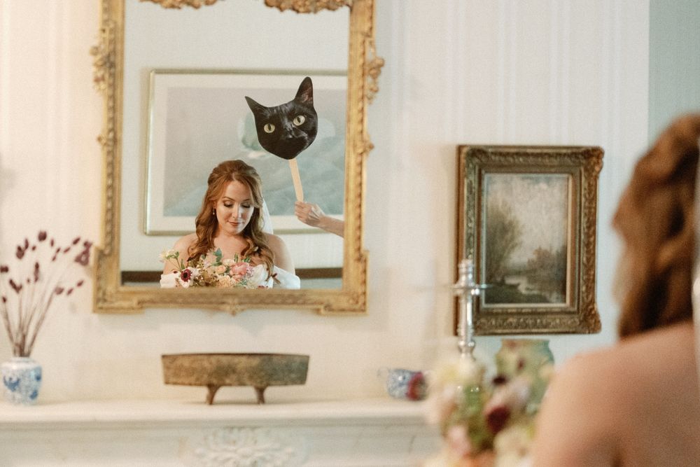 Bride holding bouquet reflected in ornate gold mirror with black cat painting above her reflection.