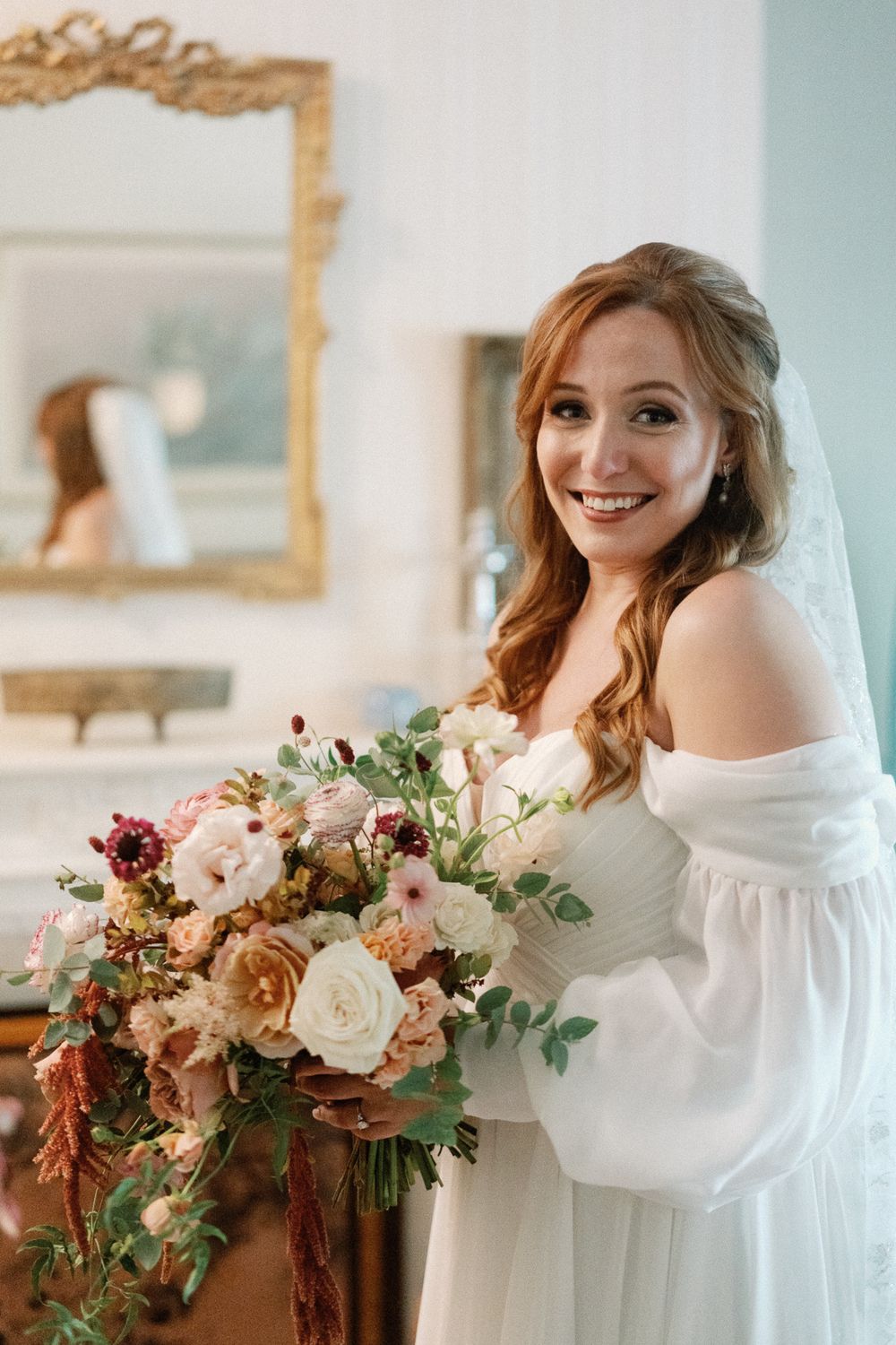 Smiling bride with red hair holding a lush bouquet of blush and burgundy flowers in an elegant white off-shoulder gown.
