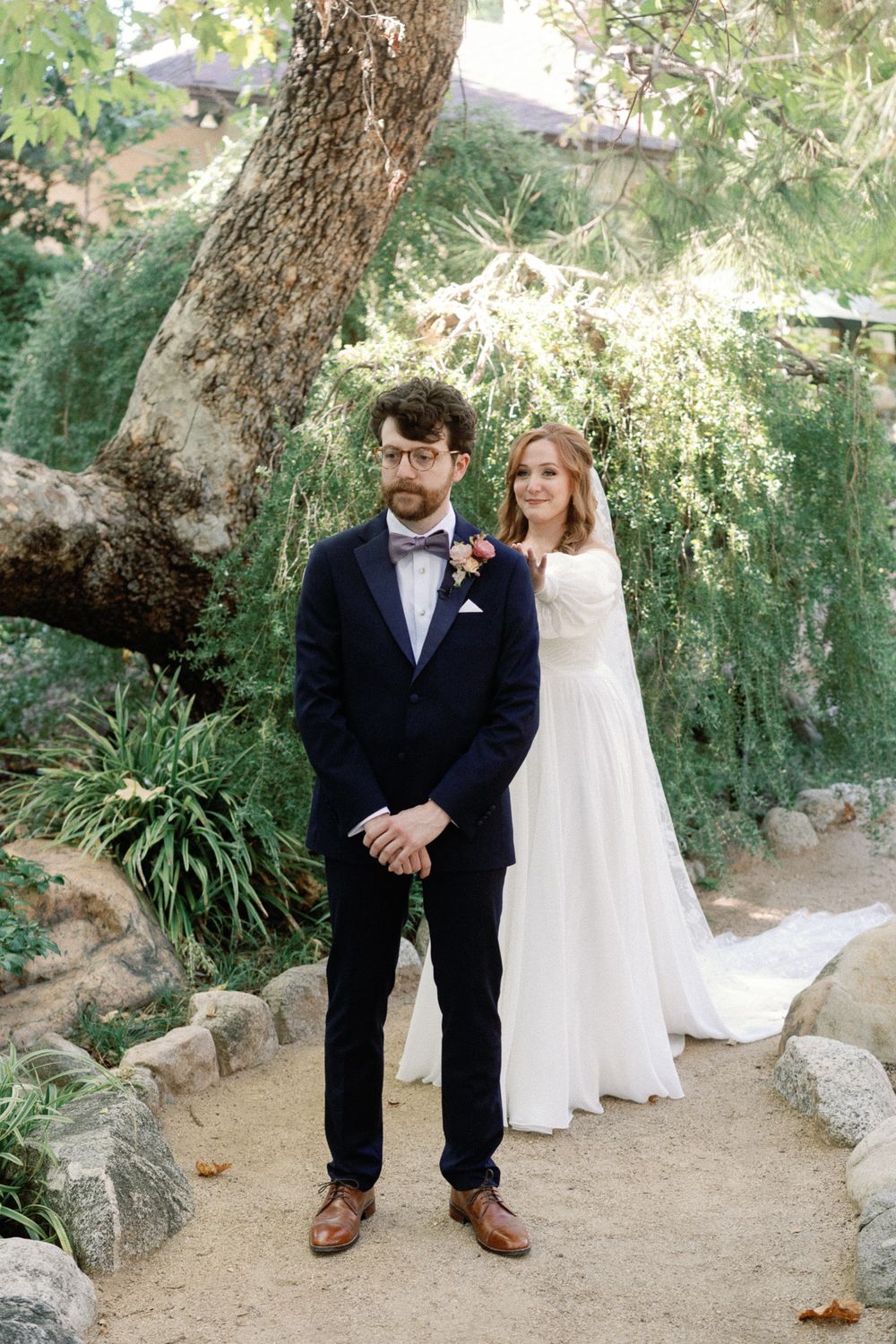 Bride and groom standing together outdoors under a large tree, groom in navy suit and bride in flowing white gown.