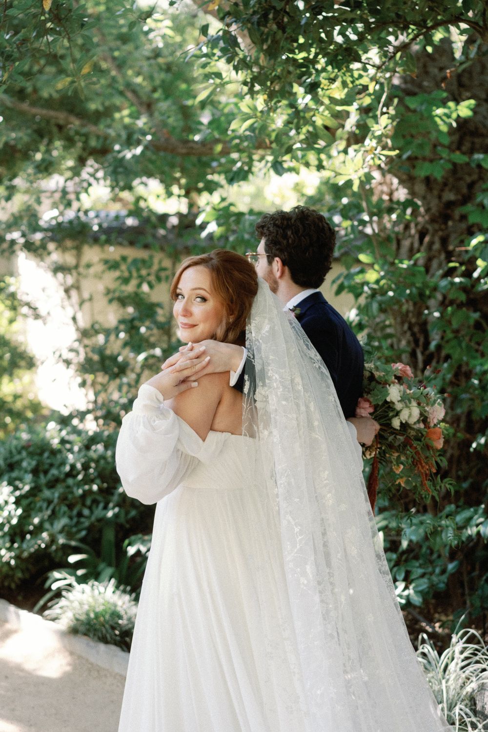 Groom embracing bride from behind in a lush garden setting, bride glancing back with a soft smile.