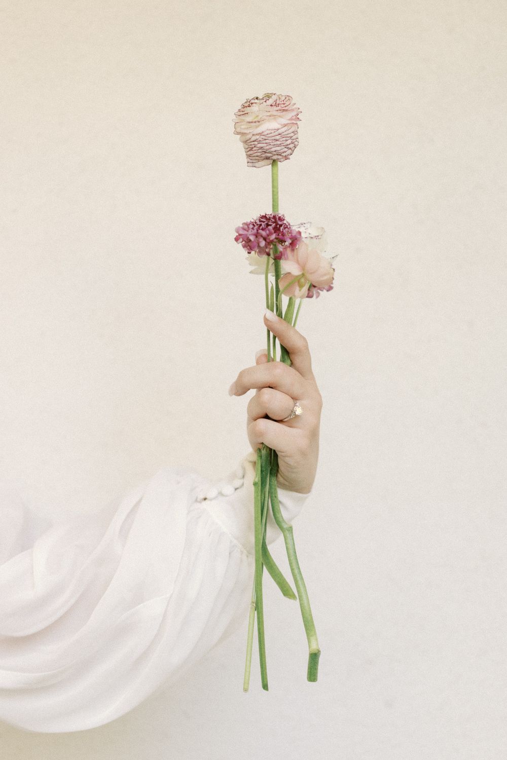 Bride holding delicate pink ranunculus and small blooms in a white lace sleeve with a diamond engagement ring.