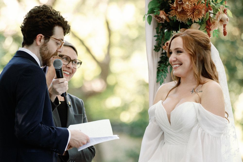 Bride smiling joyfully as groom reads vows during outdoor wedding ceremony beneath a floral arch.