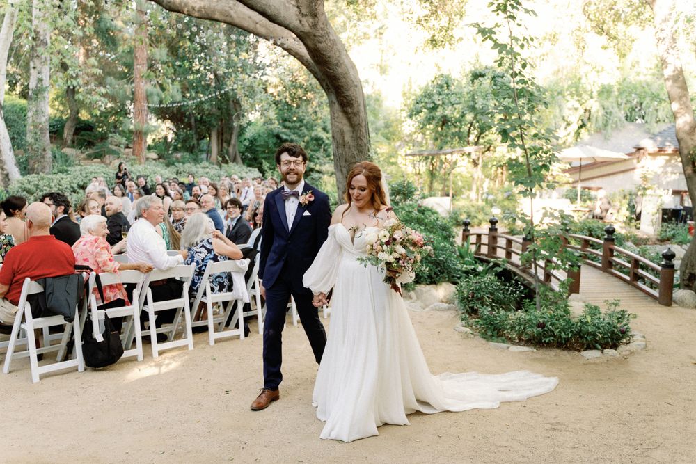 Newlyweds walk hand-in-hand down outdoor aisle at garden wedding ceremony surrounded by guests.