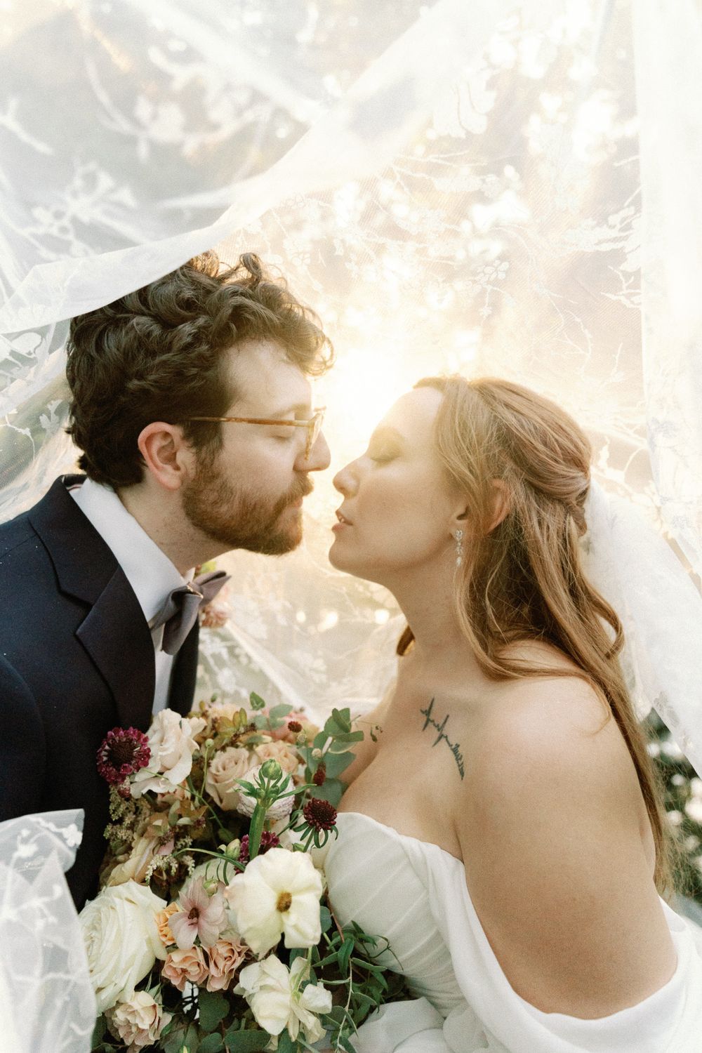 Bride and groom sharing a romantic kiss under a flowing lace veil in golden sunlight with floral bouquet.