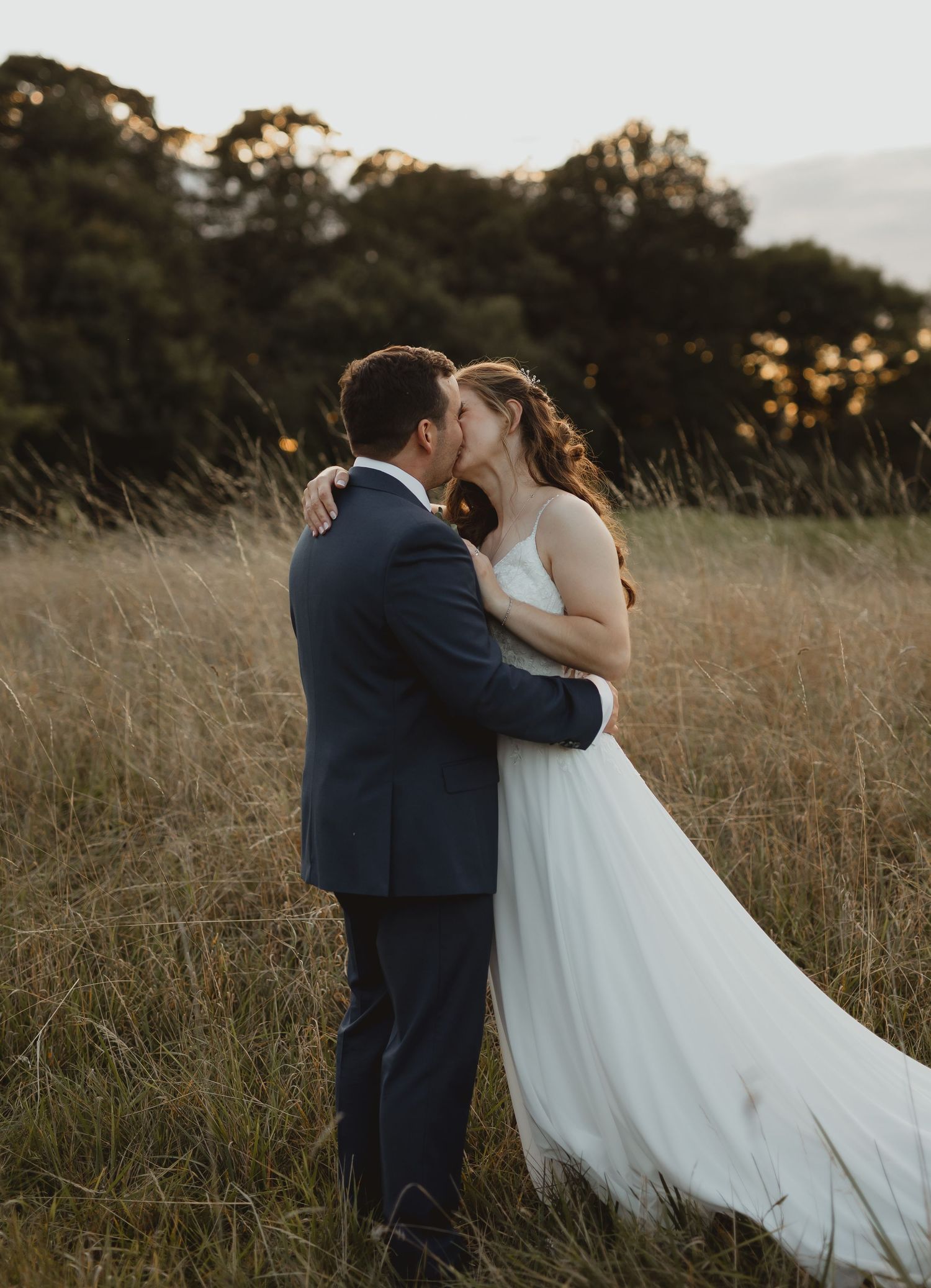 A couple embraces in a golden wheat field at sunset wearing formal wedding attire.