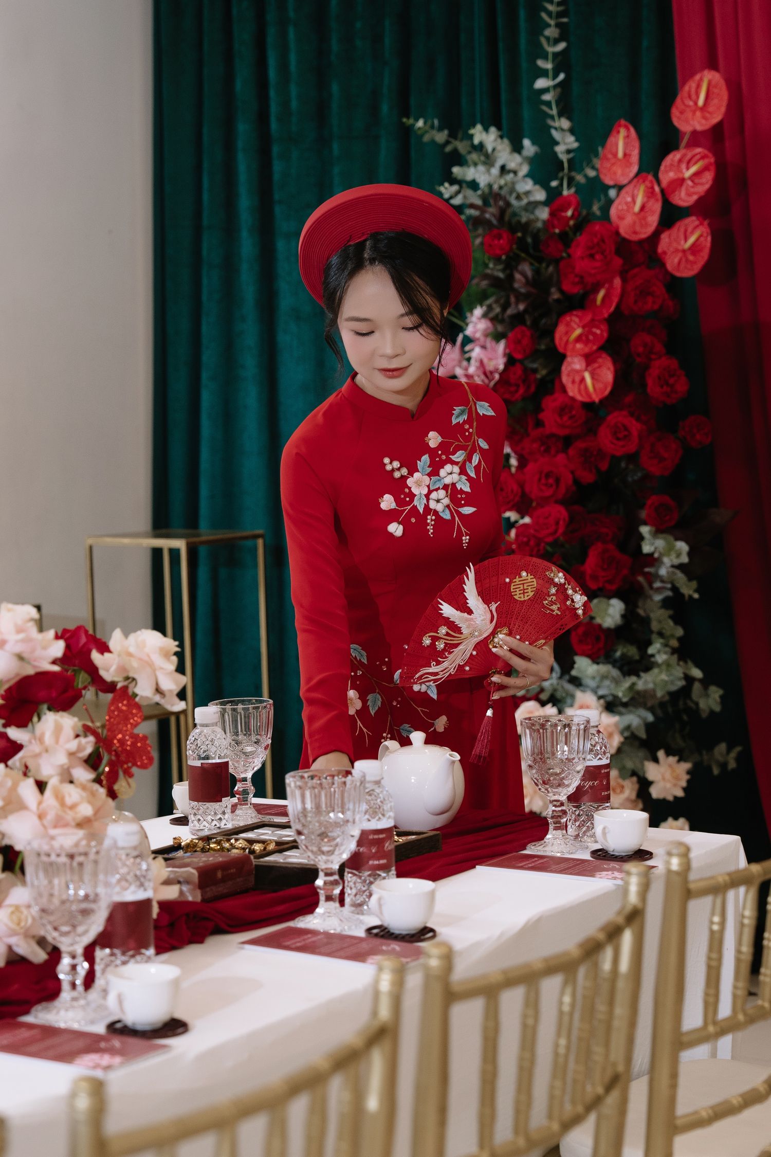 Vietnamese bride in red ao dai arranges tea ceremony table with crystal glasses and white teapot.