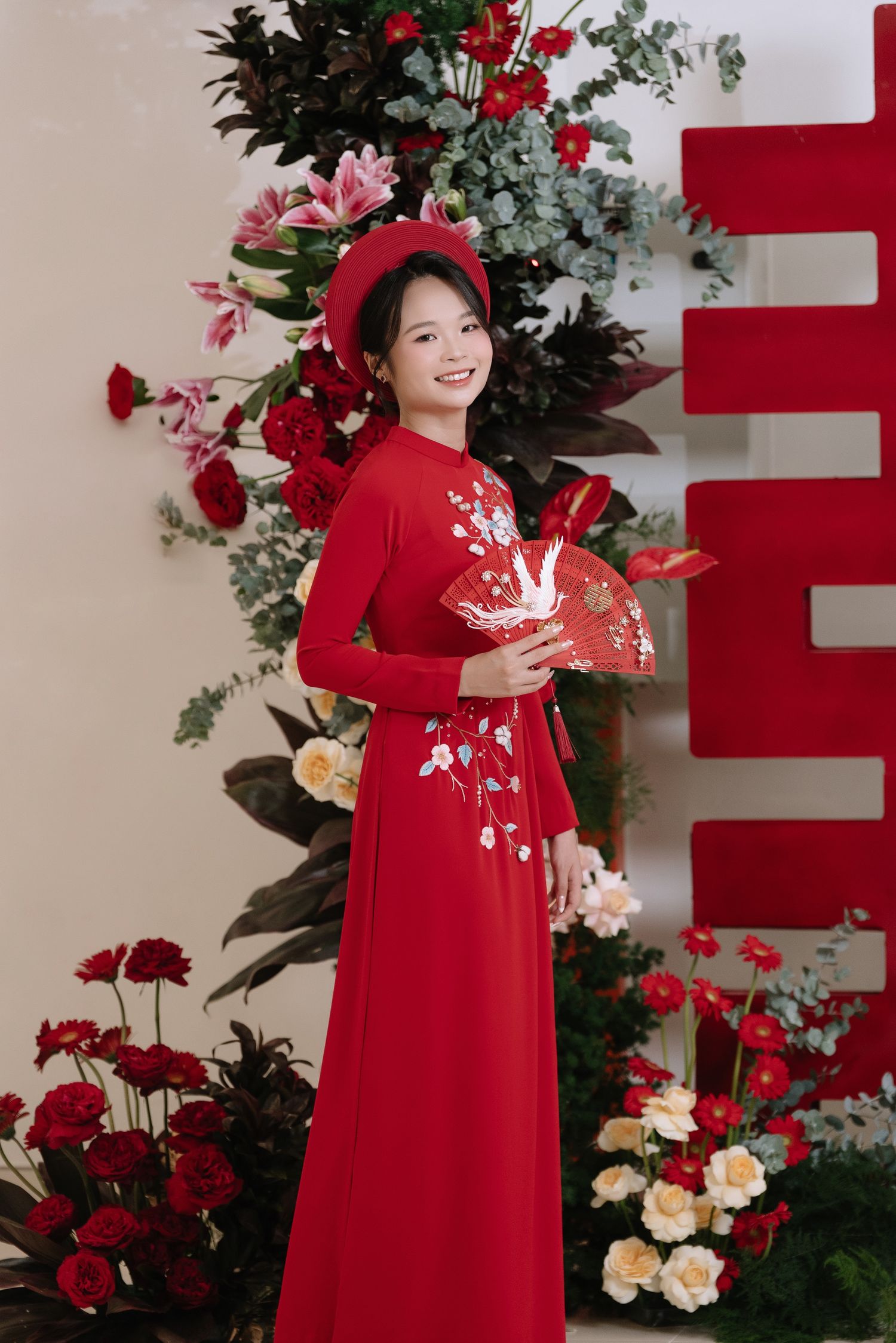 Smiling Vietnamese bride in red ao dai holding decorative fan beside floral arrangement.