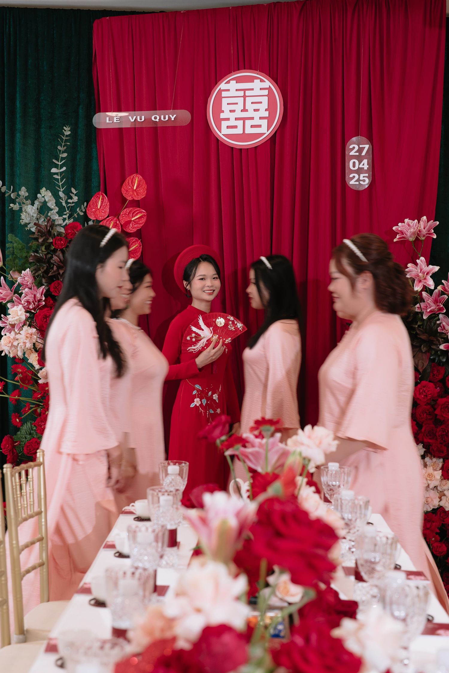 Vietnamese bride in red ao dai surrounded by bridesmaids in pink at traditional wedding ceremony backdrop.
