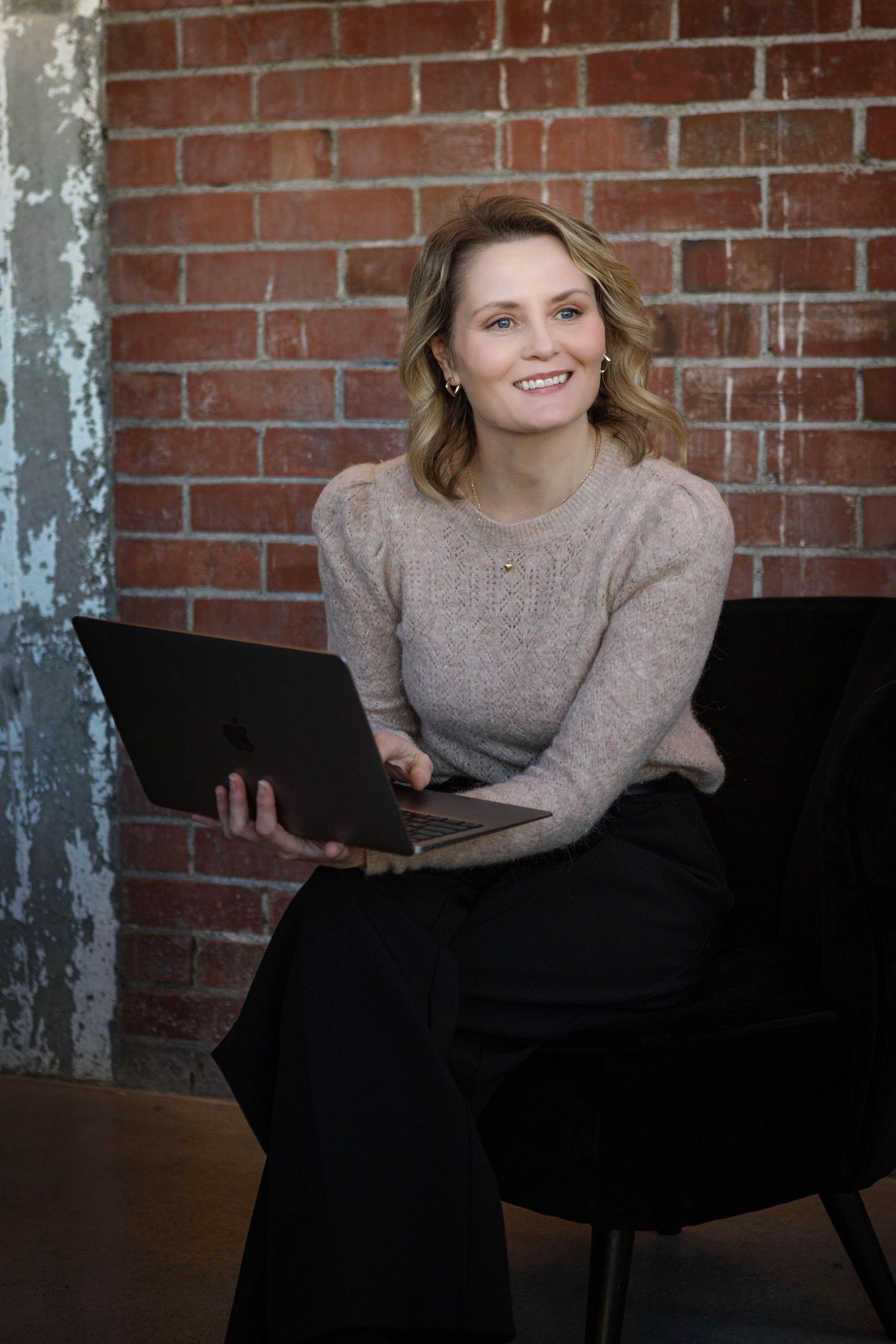 Professional seated on a black chair against a brick wall, wearing a beige sweater while working on a laptop computer.