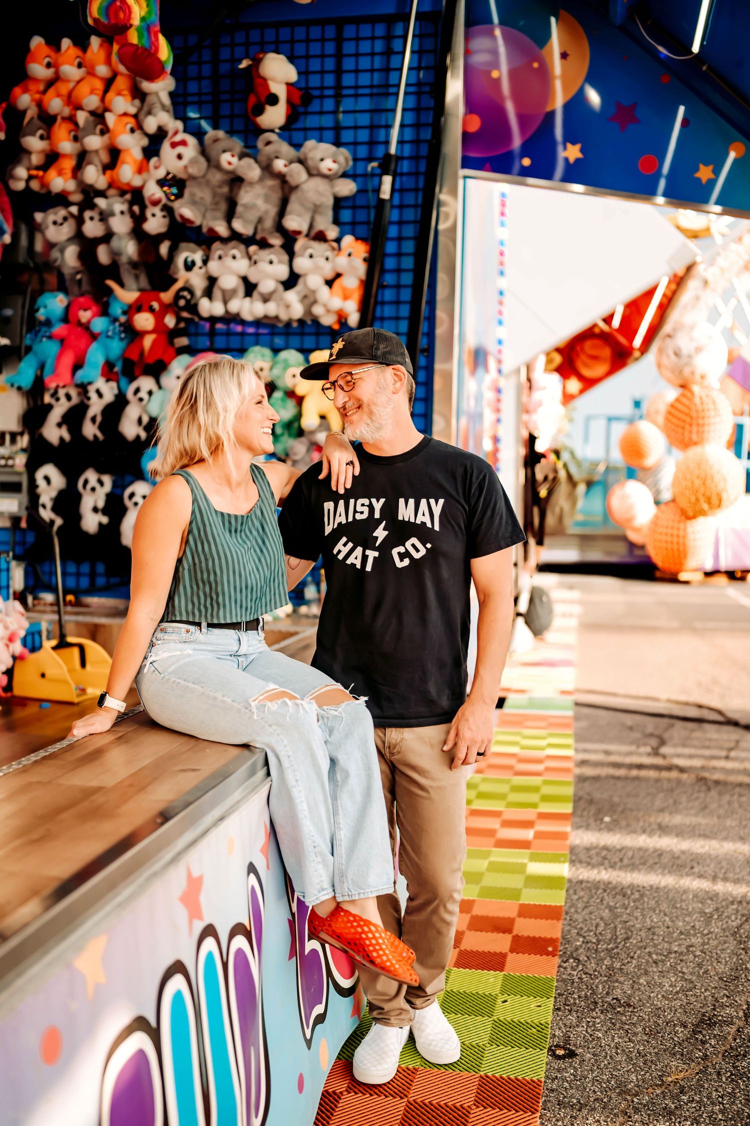 A couple sits together at a colorful carnival arcade booth with stuffed animal prizes displayed overhead.