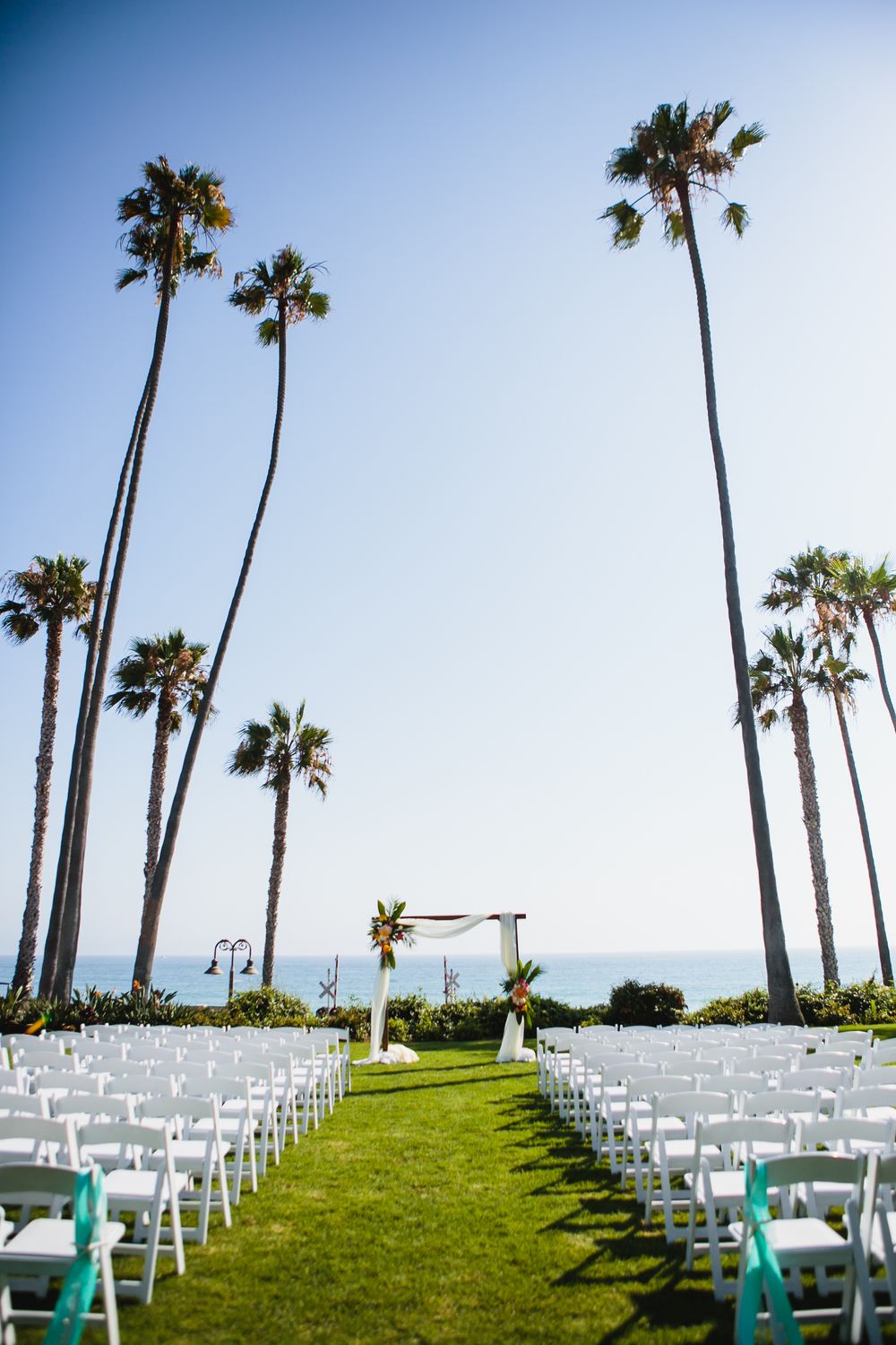 White ceremony chairs line a lush green lawn with tall palm trees and ocean views for wedding at Ole Hanson Beach Club in San Clemente.
