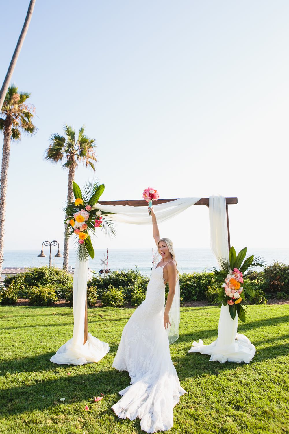 A wedding dress blows gracefully in the wind on a green oceanfront lawn with tropical palm trees in view.