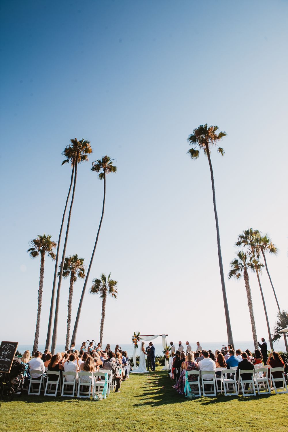 Wedding guests seated in white chairs on a grassy hilltop with tall palm trees against a bright blue sky at Ole Hanson Beach Club.