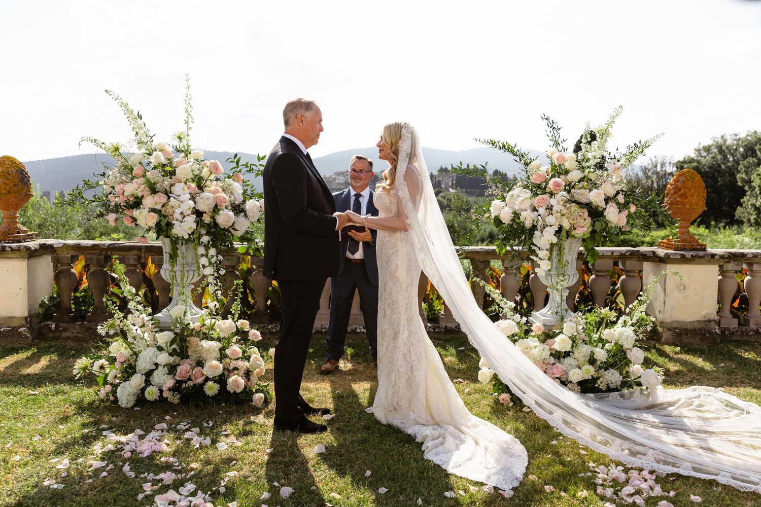 An outdoor wedding ceremony takes place on a terrace with elegant white floral arrangements and mountain views in the background.