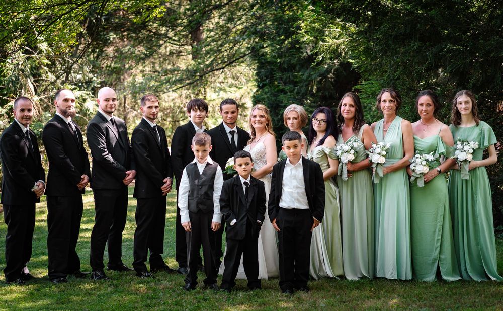 Wedding party members in black suits and sage green dresses pose together for formal group photos outdoors.