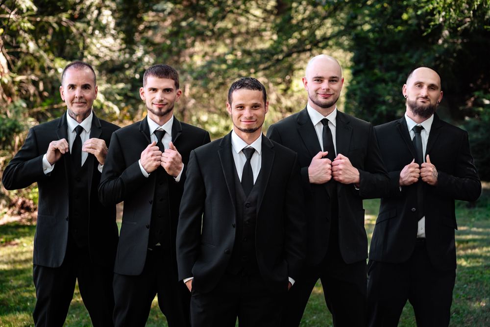 Groomsmen in black suits line up for a formal wedding photo in an outdoor setting.
