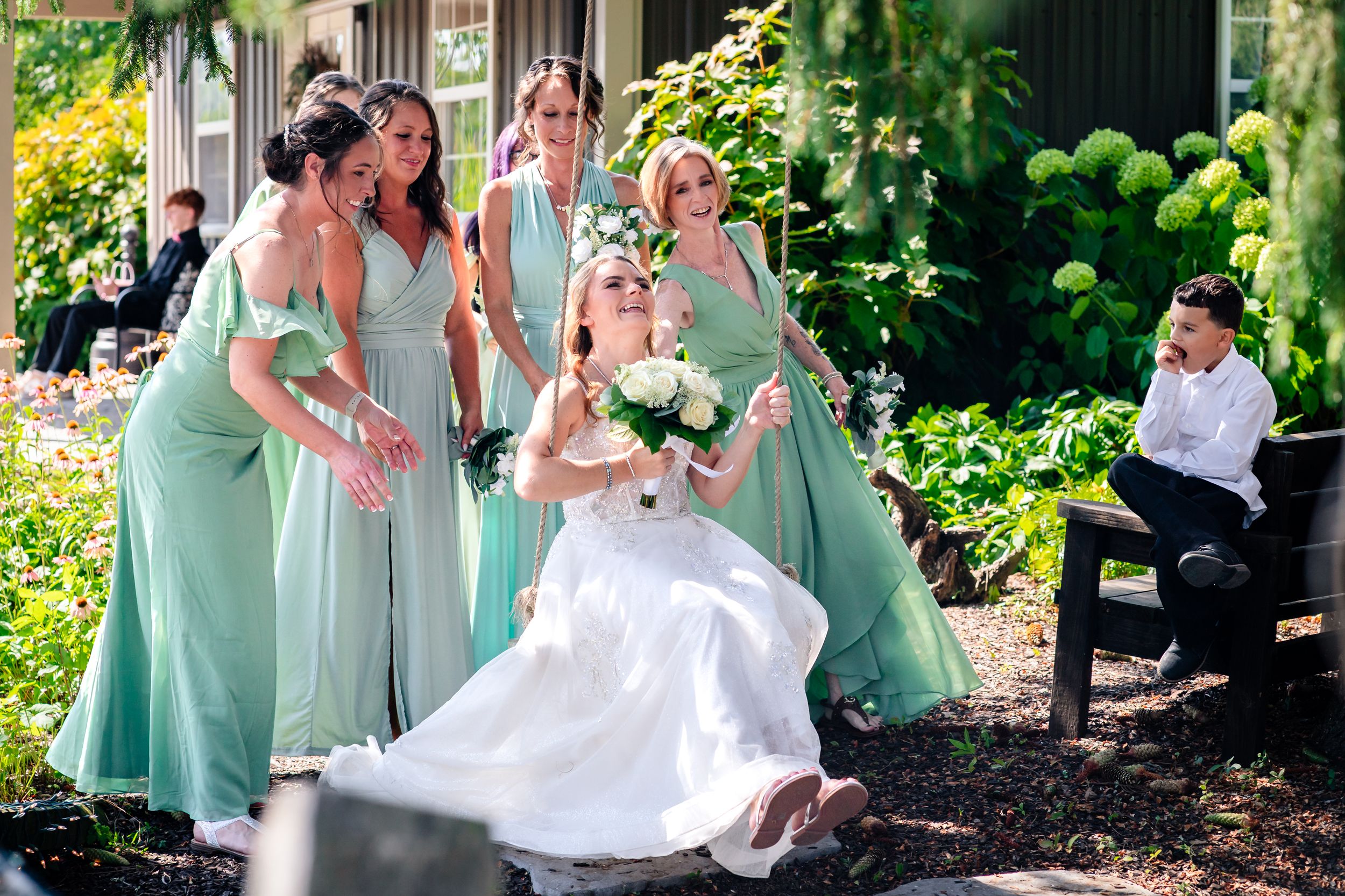 Wedding party members in sage green dresses and black suits gather for outdoor photos.